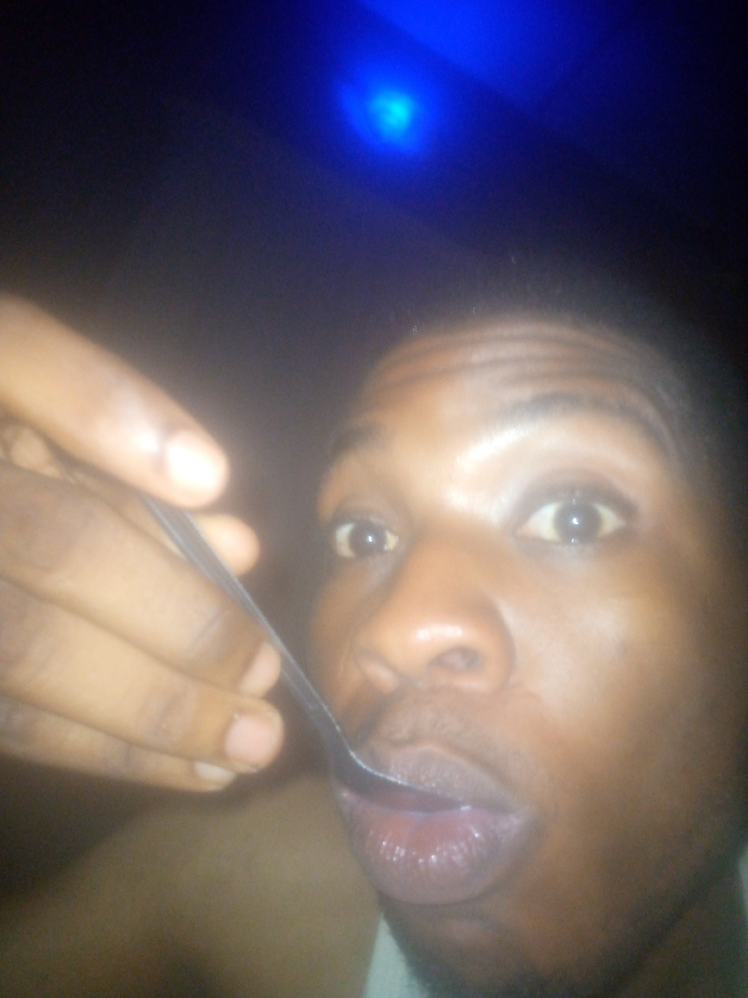 A close-up photo shows a dark-skinned person about to take food from a spoon held by another person, possibly in Unknown, Unknown. The image is poorly lit, with a blue light source in the background. The focus is primarily on the person's face and the spoon entering their mouth. The skin of both persons appears oily. The spoon looks like it is made of stainless steel. The environment is dimly lit.