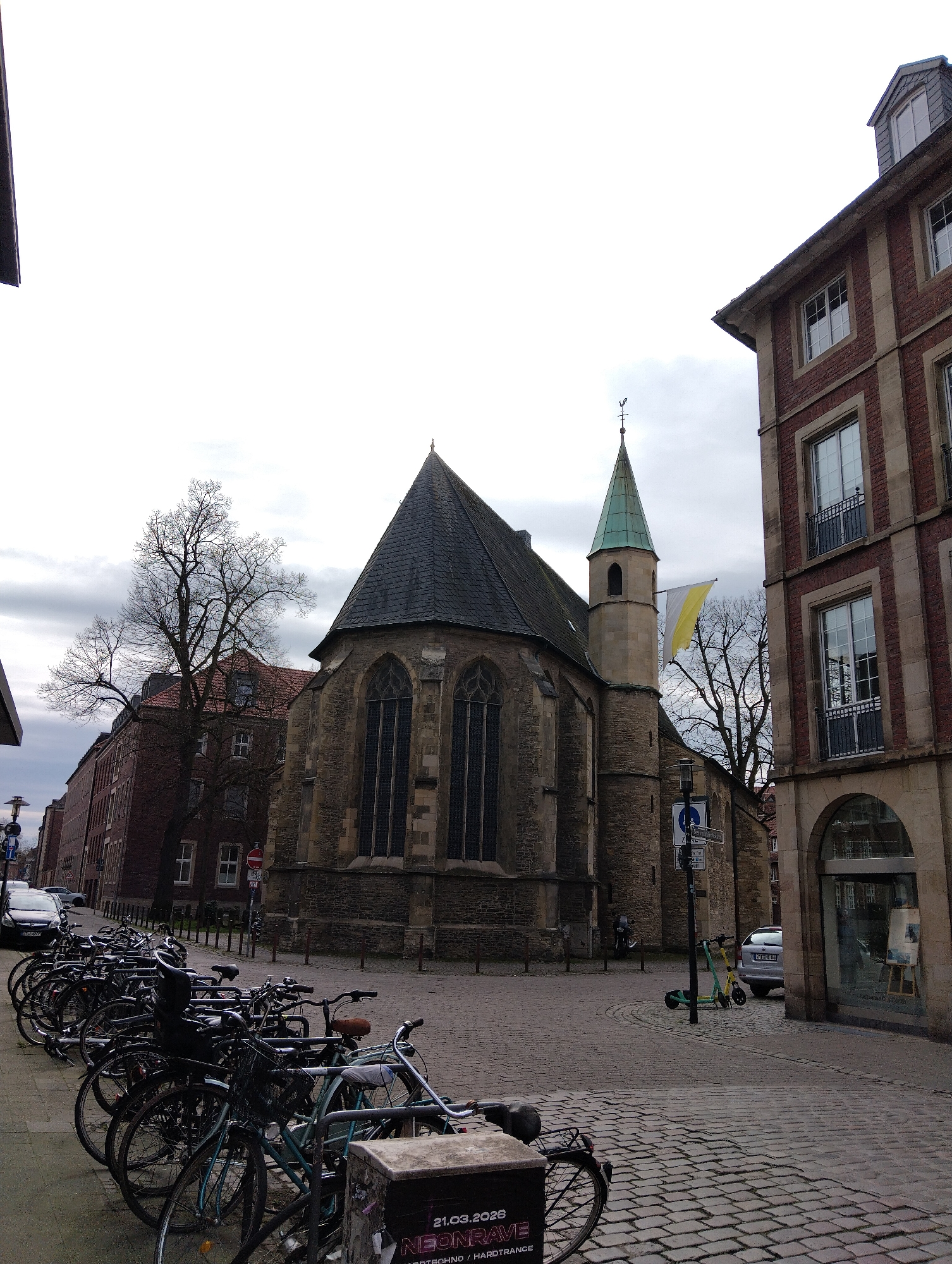 The image shows a street scene in Münster, Germany, featuring the Aegidii church prominently. The church, constructed of stone with a steeply pitched roof and a small, pointed spire, is positioned at an angle. A flag is attached to the side of the church tower. A large group of bicycles is parked to the left along the street. To the right of the church, a brick building stands with arched windows at the ground level and other windows above. The street is paved with cobblestones. Cars and scooters are parked along the street. The sky is overcast, suggesting a cloudy day.
