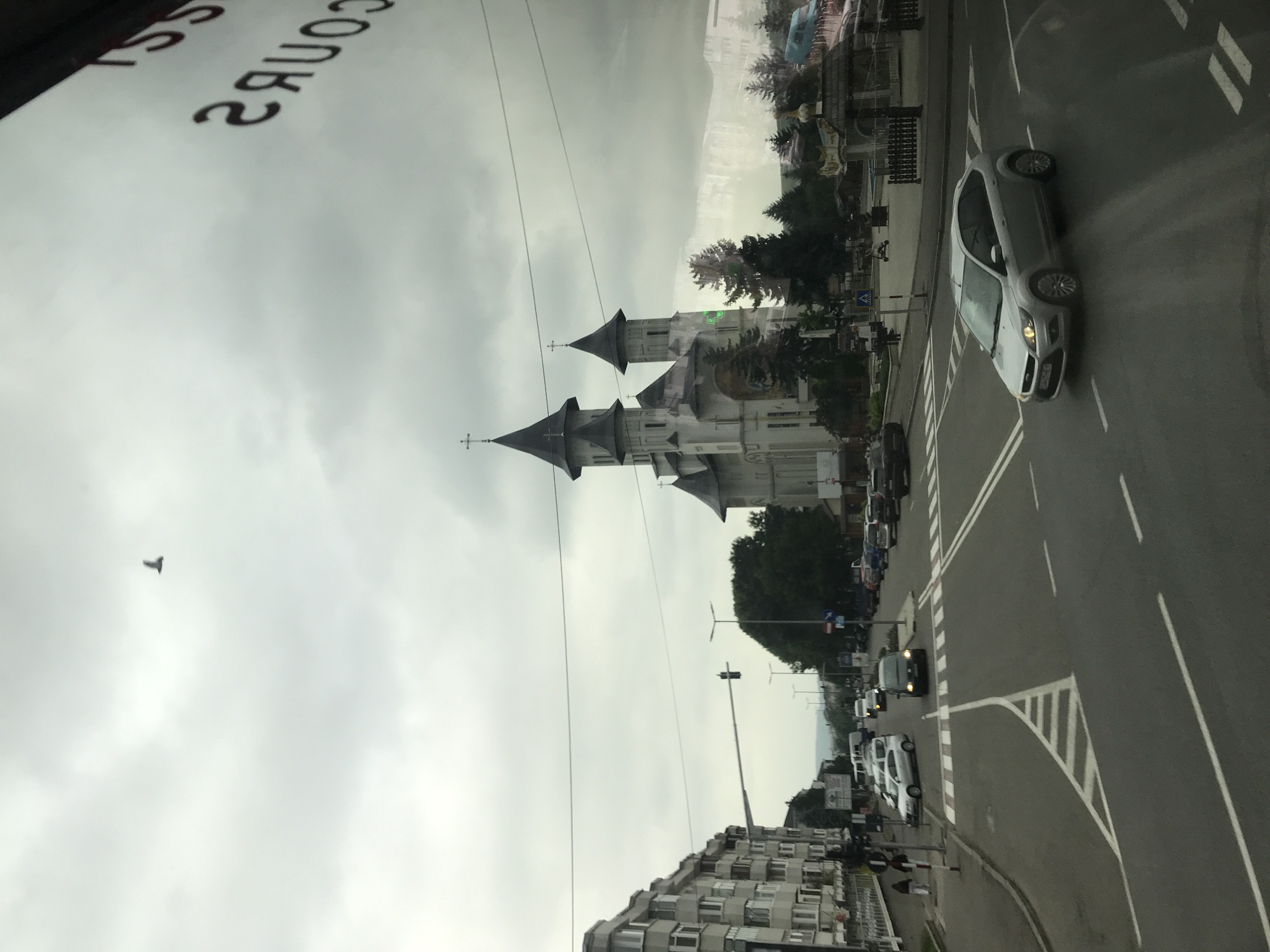 This image depicts an overcast urban street scene in Fălticeni, Romania, looking eastward along Strada Maior Ioan. The photograph appears to be taken from inside a vehicle, evidenced by the reversed text "SS COURS" reflected in the upper right quadrant of the image.
In the background, dominating the scene, stands the Neo-Byzantine style "Sfântul Gheorghe" (Saint George) Church, a prominent landmark in Fălticeni. The church is white with multiple dark, conical roofs topping its two main towers and central dome, each spire culminating in an Orthodox cross. Evergreen trees, likely firs, surround the church on a landscaped area that includes what appears to be a small public square or park with additional decorative structures, including a wooden gazebo-like feature.
In the mid-ground, a multi-lane asphalt road, identified as Strada Maior Ioan, curves through the scene. White lane markings, including a central triangular island with striped chevron markings, delineate traffic flow. A silver second-generation Ford Focus hatchback with Romanian license plates is positioned in the lower mid-ground, driving towards the right. Further down the street, an intersection is visible to the left, marked by a pedestrian crossing (zebra stripes) and a traffic light showing a green signal. Several other vehicles, including a dark minivan, are present on the road and parked along the right side.
To the left of the road, a row of multi-story (approximately 4-5 stories) residential apartment buildings with numerous balconies lines the street, indicative of mid-20th century Eastern European architecture. Two individuals, one adult and one child or smaller person, are walking on the sidewalk adjacent to these buildings near the pedestrian crossing.
The sky is uniformly overcast with light grey clouds. A lone, dark bird is captured in flight in the upper left portion of the sky. In the distant background, faint outlines of hills or mountains are visible beneath the cloudy horizon. Streetlights with modern LED fixtures are positioned along the road.