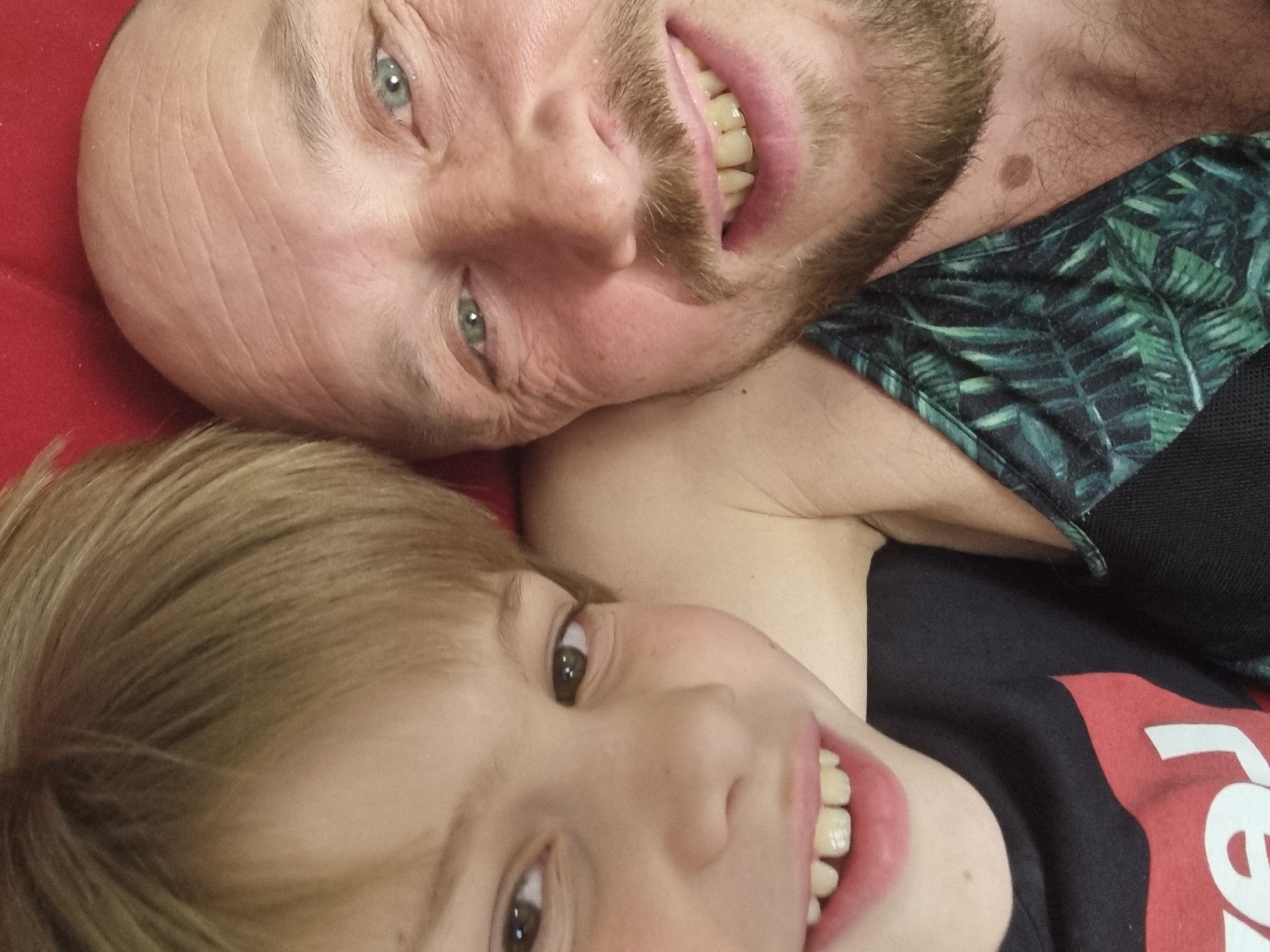 A selfie taken at close range features a balding, fair-skinned man with blue eyes and a mustache alongside a light-skinned child with brown eyes and light-colored hair. Both are smiling and appear to be lying down, with the man positioned slightly above the child. The man is wearing a garment with a green and black leaf pattern and has stubble on his neck. The child is wearing a black t-shirt with a portion of a white and red logo visible. A red surface is visible in the background. The location is unknown and there is nothing in the image to indicate it is Sochi, Russia.