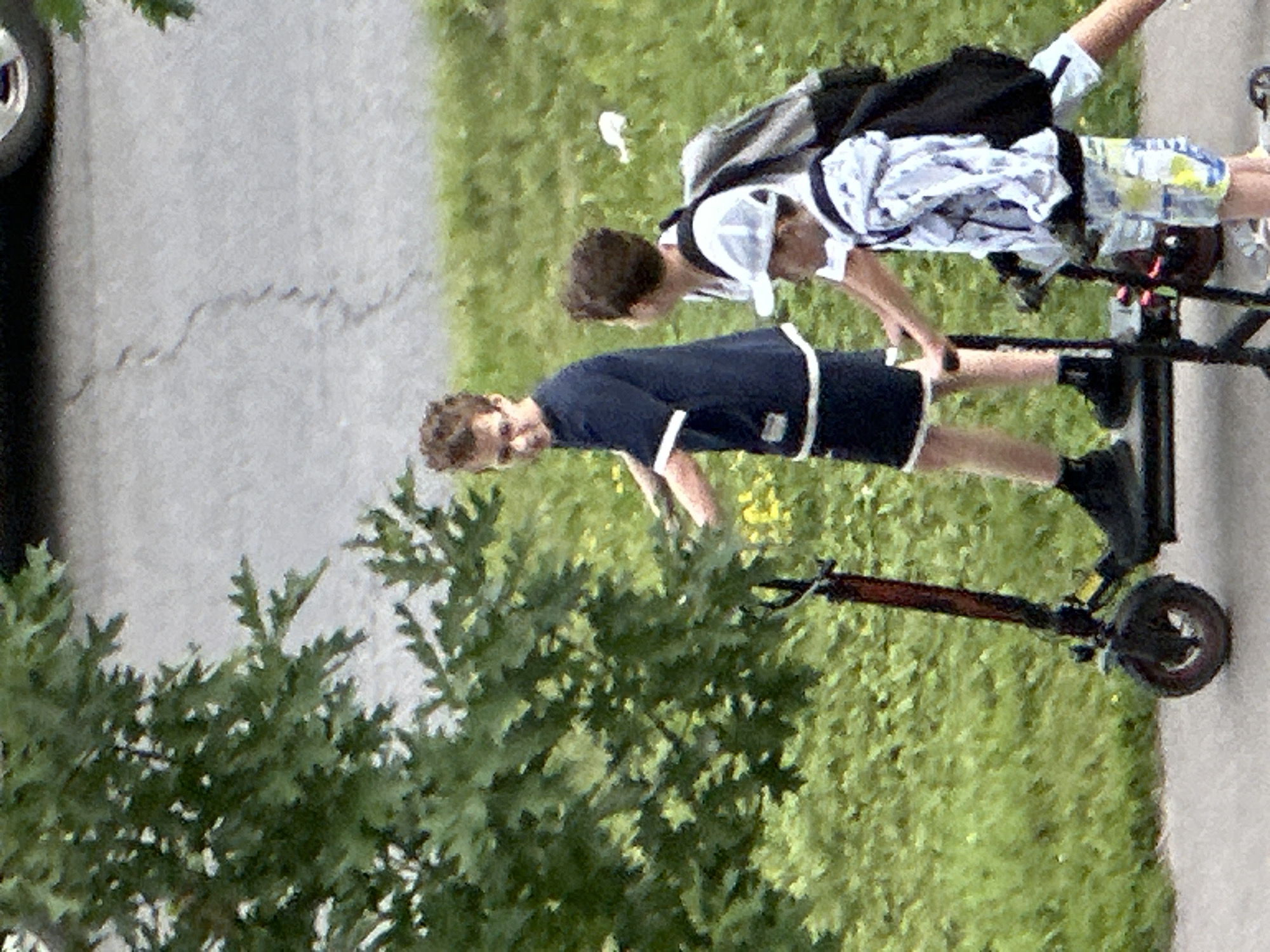 Three young individuals are present on a paved path adjacent to a grassy area with trees. The person in the foreground, on the left, is riding a black electric scooter. This individual is wearing a dark blue t-shirt with white trim on the collar and sleeves, dark shorts, and black shoes. Their hands are on the handlebars of the scooter.
To the right and slightly behind, a second individual is visible from the chest up. They are wearing a white t-shirt and a white baseball cap. This person appears to be looking towards the right.
Behind the second individual, a third person is seen from the waist up. They are wearing a light-colored hooded jacket with a patterned design, dark shorts, and are carrying a black backpack. They are also wearing a white baseball cap.
The environment consists of a paved pathway, a green grassy area, and a tree with green leaves on the left side of the frame. A hint of a vehicle tire is visible at the top of the image, suggesting it is on a road or parking area. The scene appears to be outdoors during daylight. The image is from Podolsk, Russia.