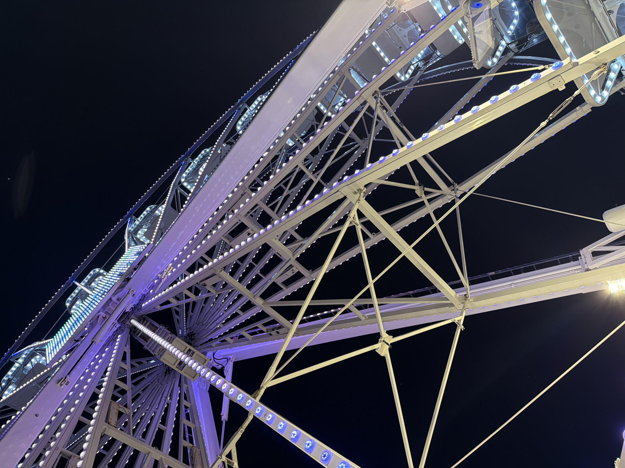 The image shows a close-up, low-angle view of a Ferris wheel at night, illuminated by numerous white and purple lights. The wheel's structure is intricate, composed of white metal beams and supports. The sky is dark, indicating nighttime. The Ferris wheel is likely located in the city of Gatteo, Italy, as per the prompt's context. No people are clearly visible in the frame. The perspective emphasizes the scale and illumination of the amusement ride against the dark sky.