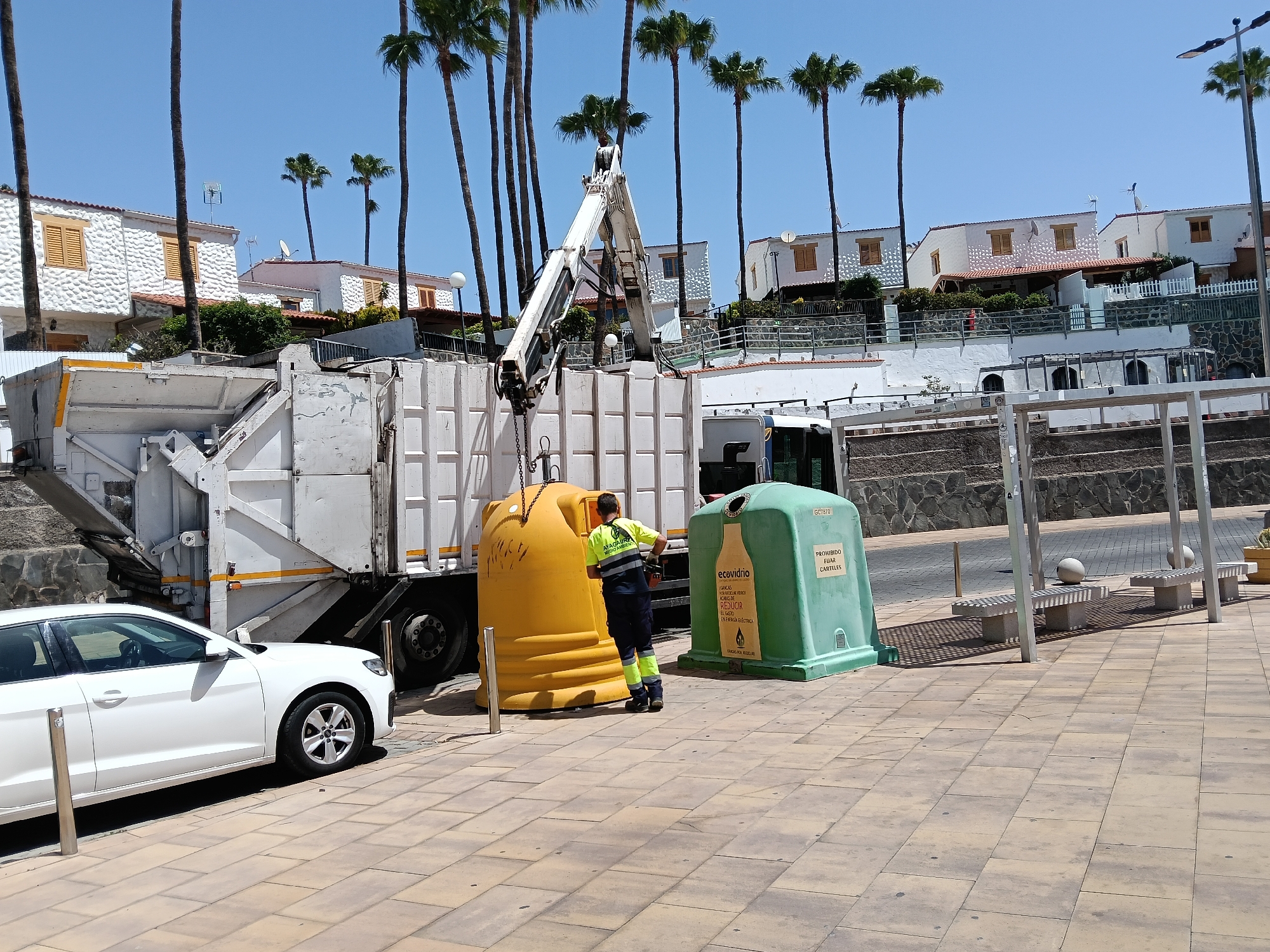 This bright, sunny image, captured in San Bartolomé de Tirajana, Spain, shows a waste collection operation in progress. A garbage truck, predominantly white, is actively emptying a large yellow communal recycling bin. The truck's automated crane arm is extended, lifting the yellow bin upside down into the truck's rear compartment. A waste management worker, wearing a high-visibility yellow and dark blue uniform with "NAGAIBA MEDIO AMBIENTE" on the back, stands near the yellow bin, supervising the process.
To the right of the worker is a green, bell-shaped communal recycling bin specifically for glass. Visible text on this bin includes "GCT870", the brand "ecovidrio", and instructions such as "GANCHOS PARA VOLCAR VIDRIO" (hooks for emptying glass), "ACABAS DE REDUCIR EL GASTO EN ENERGÍA ELÉCTRICA" (you have just reduced the expenditure on electrical energy), and "PROHIBIDO FIJAR CARTELES" (prohibited to affix posters), followed by "PARA RECICLAR" (for recycling).
In the foreground, a white sedan is parked on the light-colored paved sidewalk. The background is dominated by a clear, azure sky and numerous tall palm trees. Beneath the palm trees, a row of white residential houses with terracotta-colored roofs and prominent wooden shutters are visible, built on a gentle slope. Further to the right, beyond the recycling bins, white concrete benches are situated under a modern shelter structure. The scene takes place during the day under bright sunlight, indicated by crisp shadows.