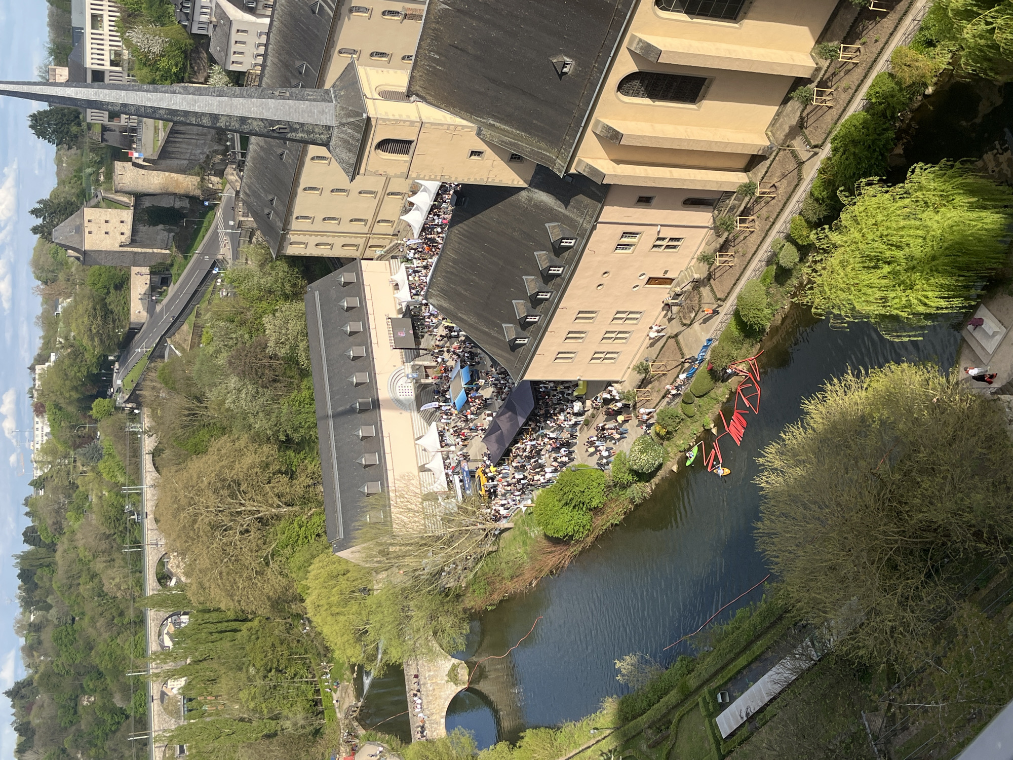 The image shows an aerial view of a densely populated outdoor event held along the banks of the Alzette River in Luxembourg City, Luxembourg. Numerous people are gathered at tables and in open areas, suggesting a festival or market. Buildings, including a prominent church with a spire, surround the event. The river flows past the gathering, and several bridges are visible in the background, with pedestrians walking across them. Mature trees line the riverbank, contributing to the urban green space. Several canoes/kayaks are docked along the river.