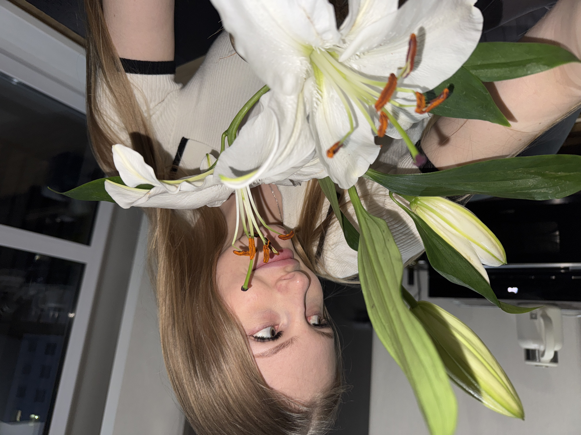 A woman with light brown hair, wearing a cream-colored sweater with a black stripe on the sleeve, is holding a white lily flower up to her face. The lily's stamen are touching her lips. She is looking to the right of the frame. The background includes a window showing a night scene of a city. The picture was likely taken in Perm, Russia.