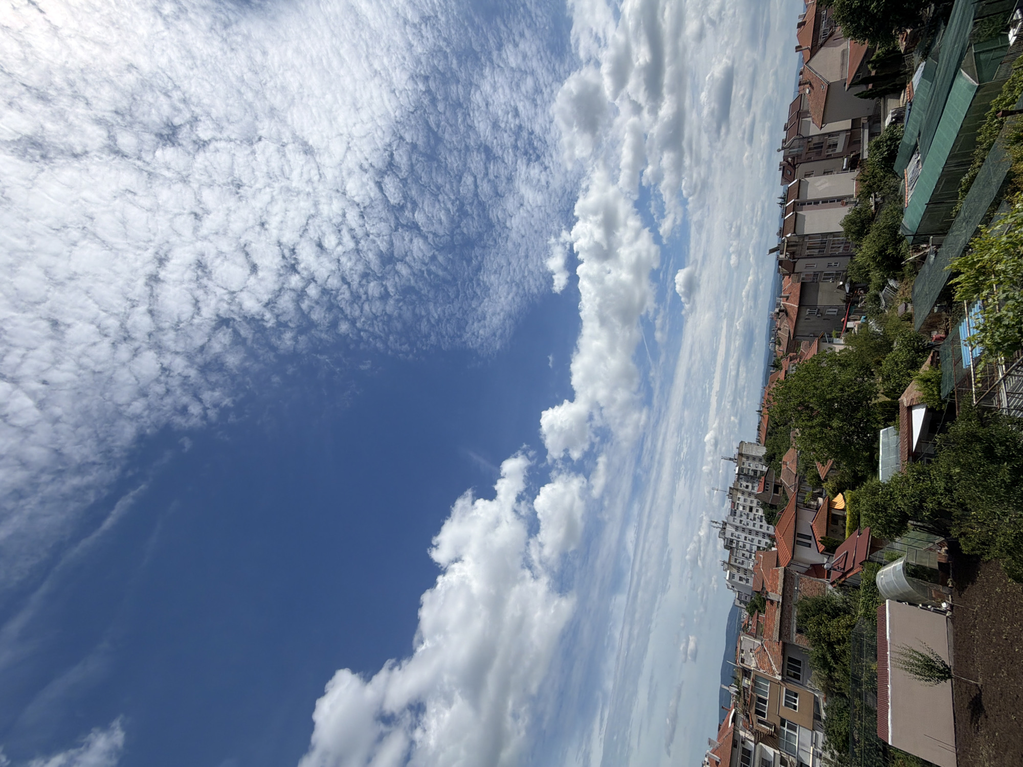 The image shows a view of the city of Burgas, Bulgaria, on a clear day with scattered clouds. The foreground consists of a garden area with soil and some small trees. In the middle ground, there are several residential buildings with red-tiled roofs, interspersed with green trees. Some of these buildings appear to be older houses, while others are larger apartment complexes. Several greenhouses are visible in the garden area. In the background, a large apartment building stands on a hill, and the cityscape extends towards the horizon. The sky is a vibrant blue, with a mix of large cumulus clouds and smaller, wispy cirrus clouds that create a textured pattern across the upper portion of the image. There are no people or animals visible in the image. The scene appears to be a typical residential neighborhood in Burgas, likely captured from an elevated viewpoint.