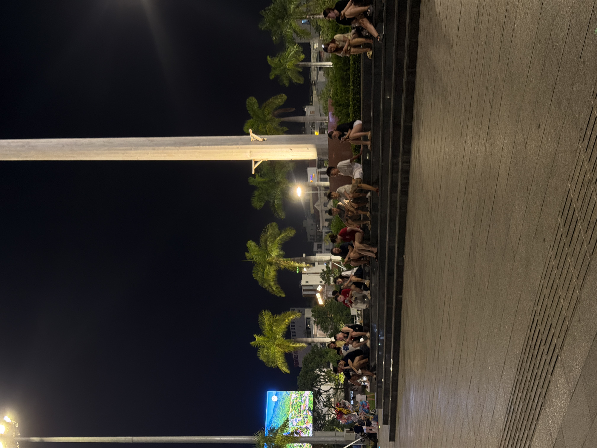 A group of young adults are sitting on dark, wide stone steps in what appears to be a public square at night. The scene is illuminated by large overhead lights and a brightly lit digital screen displaying a landscape image of mountains and a moon. Palm trees are scattered throughout the background, and buildings can be seen behind them. Some individuals are looking at their phones, while others are conversing. The steps are part of a paved area that leads to the steps, with textured concrete in the foreground. The overall atmosphere suggests a relaxed evening gathering in an urban setting. This location is likely the Bach Dang street pedestrian street in Da Nang, Vietnam, known for its scenic riverfront and evening activities.