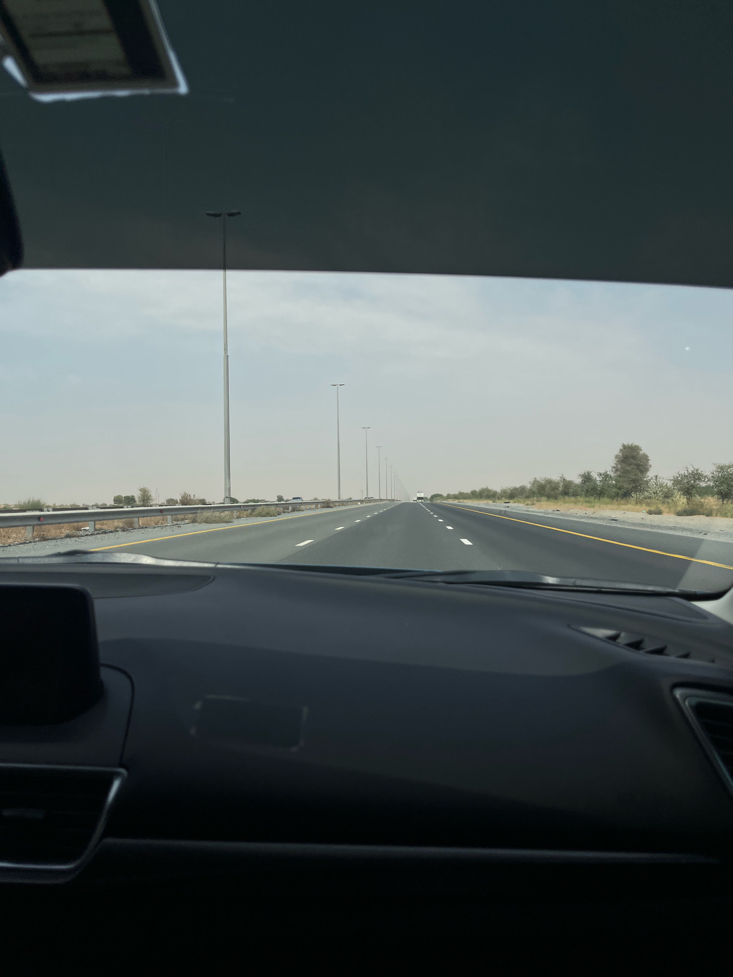The image is taken from inside a car, looking forward through the windshield. The car is on a multi-lane highway that stretches into the distance, flanked by desert vegetation and streetlights. The sky is overcast and hazy, suggesting a warm climate. The dashboard of the car is visible in the foreground, with a dark, matte finish. The highway appears to be in an open, rural area, possibly on the outskirts of a city. There are no visible people in the image. The location is identified as Mohadhub, United Arab Emirates.