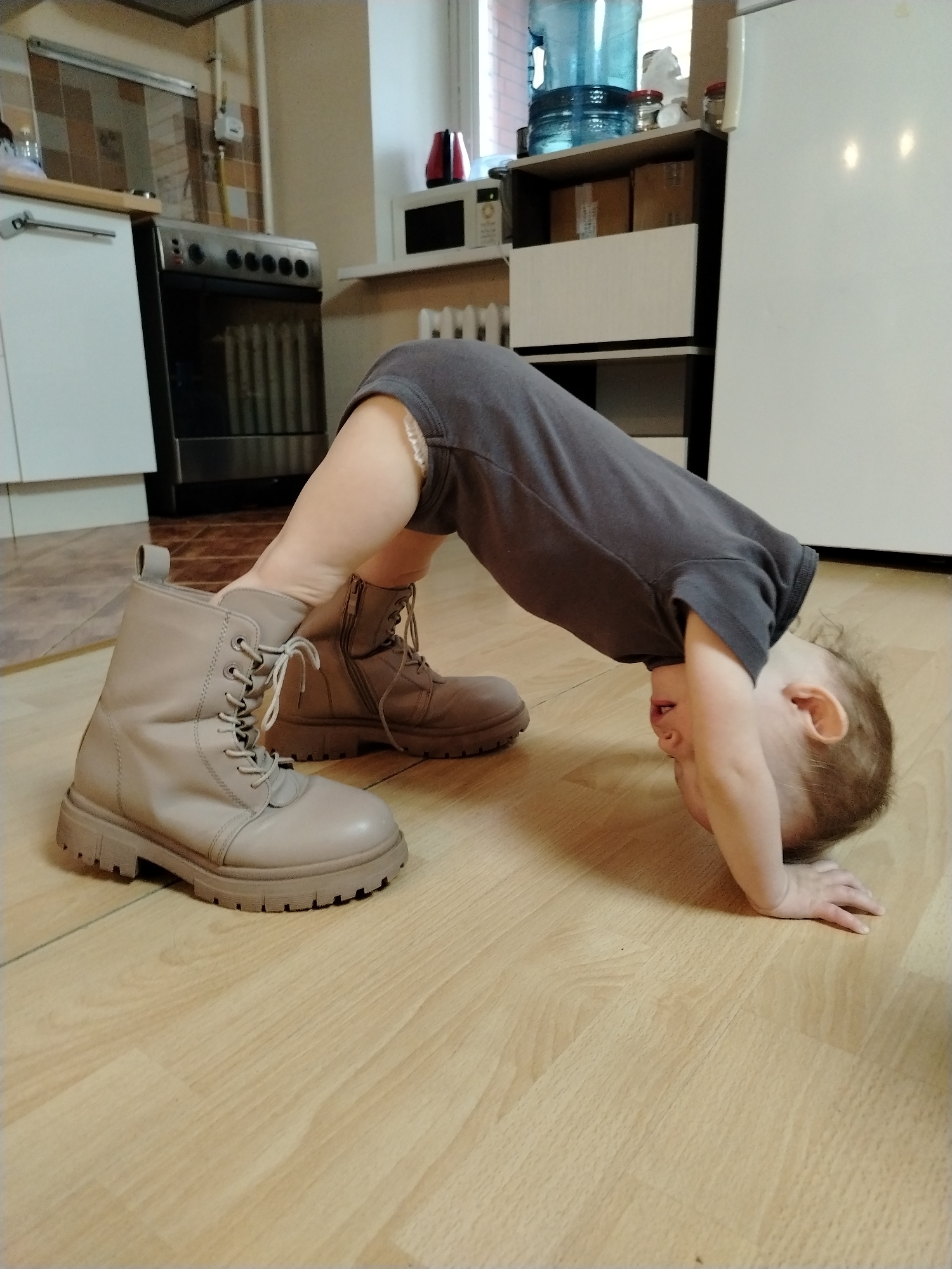 The photo shows a toddler wearing oversized boots in a kitchen setting. The child, dressed in a gray onesie, is bent over, with its head near the floor and its legs extended into the boots. The boots are light brown leather lace-up combat boots. The floor is light wood-grained laminate.
In the background, there is a white kitchen cabinet, a black stove, and a microwave. There is also a water cooler on top of a shelf.
The location is Chaikovsky, Russia, based on visual cues in the background suggestive of the region.