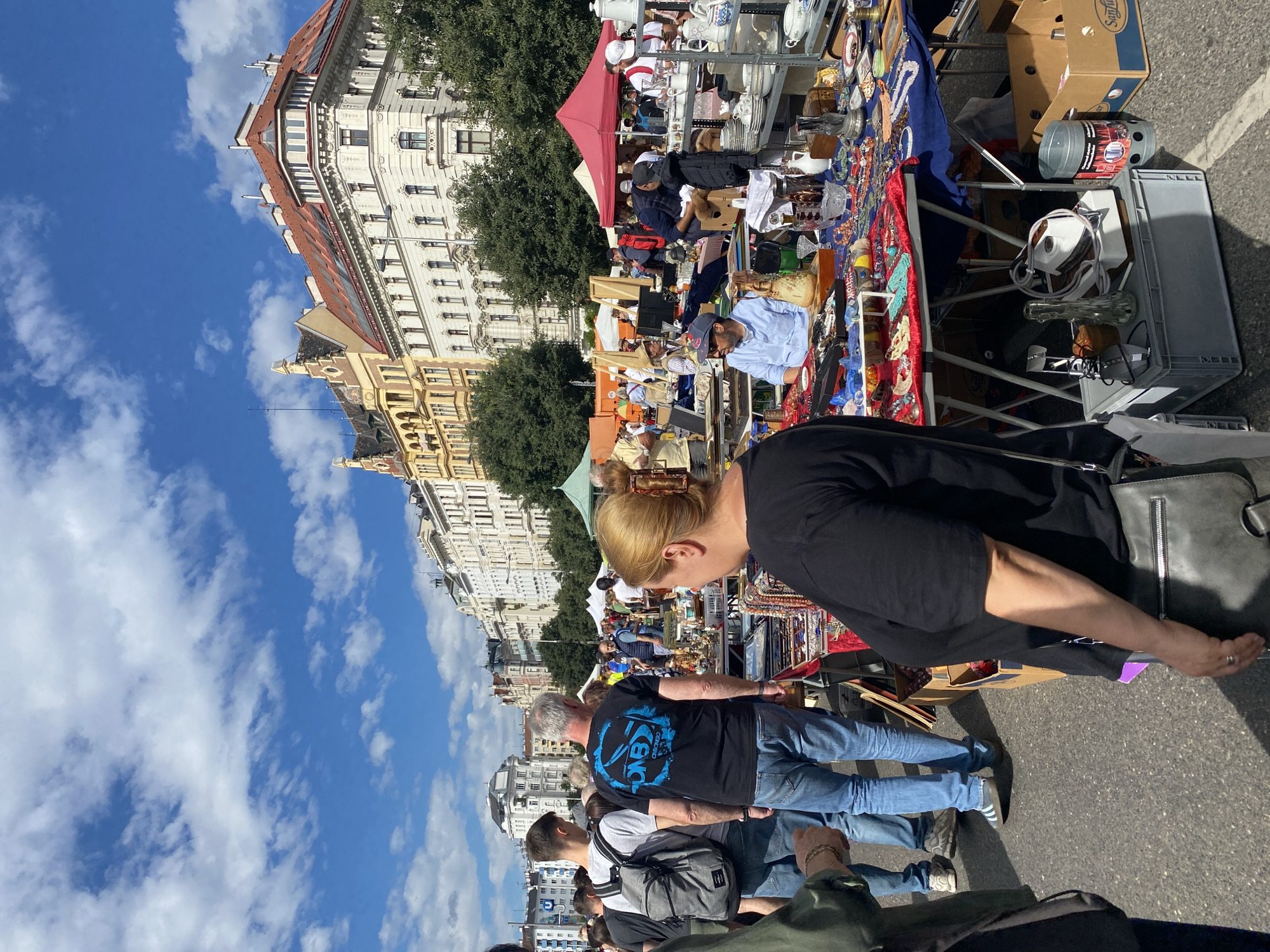 The image captures a vibrant outdoor market scene in Vienna, Austria, likely a flea market, with stalls laden with various items for sale. In the foreground, a woman with blonde hair tied up is seen from the back, wearing a black t-shirt and a black shoulder bag. She is looking towards a table displaying an assortment of jewelry and trinkets. Behind her, two men are walking away from the camera; one wearing a black t-shirt with a blue graphic and jeans, the other in a grey t-shirt, jeans, and a backpack.
The market extends into the background, featuring more tables with a diverse array of goods, including pottery, glassware, and decorative items. Several tents, one prominently red, are set up to provide shade. People of various ages are browsing the stalls. The backdrop is dominated by the distinctive architecture of Vienna, with multi-story buildings featuring ornate facades and red-tiled roofs, typical of the city's historic districts. The sky is bright blue with scattered white clouds, indicating a pleasant day. The ground is paved with asphalt.