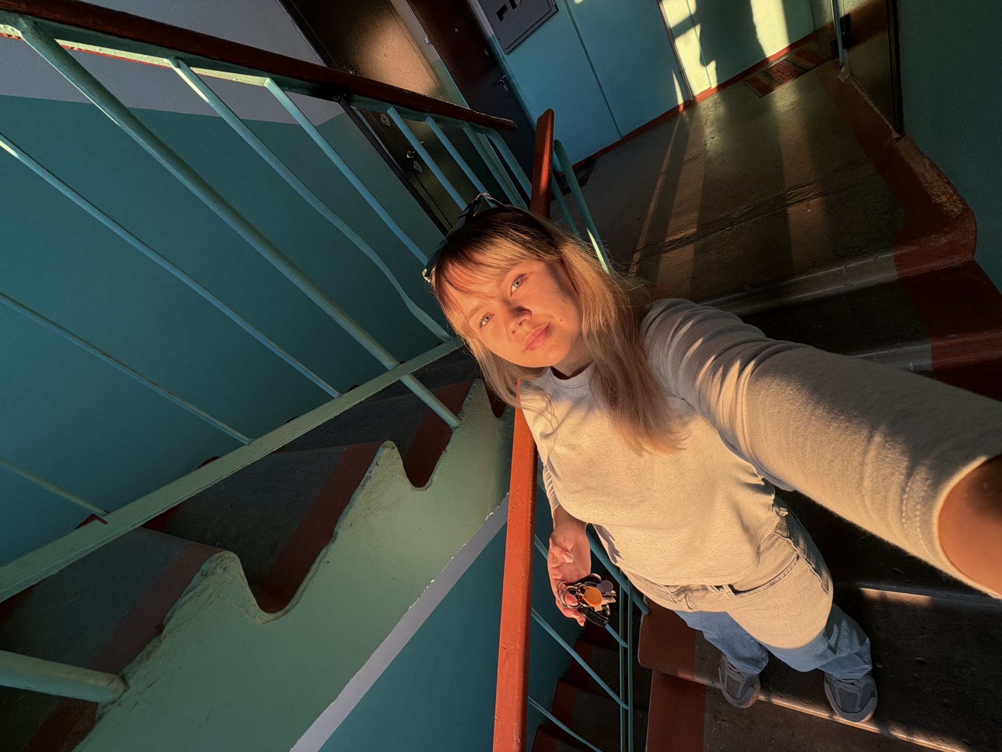 A young woman is captured in a high-angle selfie within an indoor stairwell in Chaikovsky, Russia. She has blonde hair with bangs, fair skin, and is looking directly at the camera with a calm, pleasant expression, featuring a slight smile. She wears a light-colored long-sleeved top, light-colored jeans, and dark sneakers. Sunglasses are perched on top of her head. In her right hand, she holds a bunch of keys with several colorful keychains.
The stairwell features a prominent railing system painted in a teal or turquoise color, with an orange-red handrail running along the top. The stairs themselves are dark-colored, possibly painted dark brown or black, with orange-red accents along their edges. The walls of the stairwell are also painted in a similar teal/turquoise hue.
Strong, golden sunlight streams into the stairwell from an unseen window or opening, casting long, dramatic shadows across the stairs and walls, creating a warm and somewhat atmospheric glow. The light illuminates parts of the woman's face, arm, and clothing, highlighting the textures of the stairwell. The overall design of the stairwell appears robust and functional, characteristic of older residential or public buildings. No visible text is present in the image.