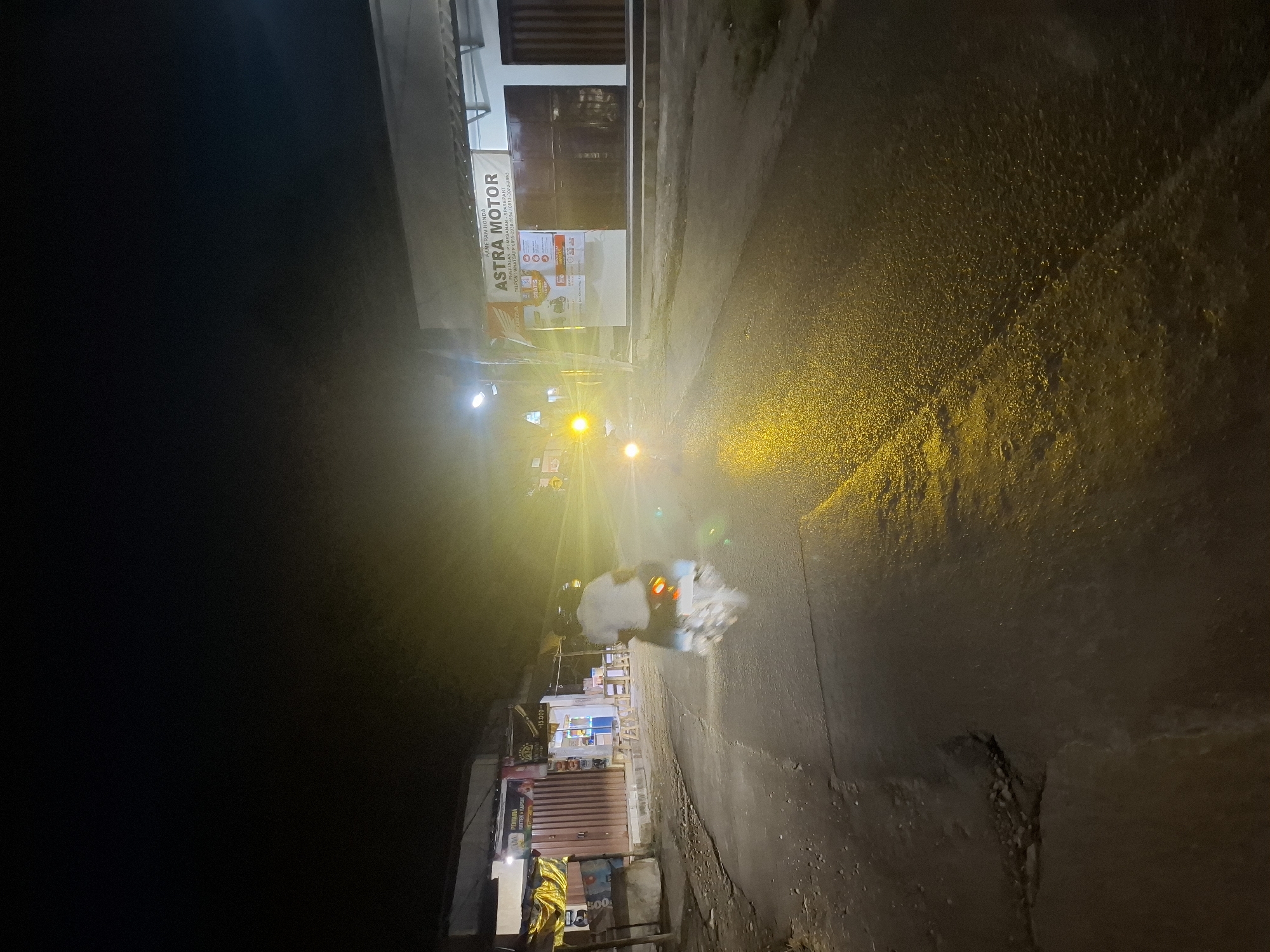In the city of Ciheulang Tonggoh, Indonesia, at night, a person is riding a motorcycle down a paved road. The motorcycle's headlights illuminate the wet asphalt, reflecting the bright yellow light. To the right of the road, a building with a banner reading "ASTRA MOTOR" is visible, suggesting it's a motorcycle dealership or repair shop. The building has a white facade and dark brown doors. To the left, another building with corrugated metal siding and a yellow tarp is present, appearing to be a storefront. The overall scene is illuminated by streetlights and the motorcycle's headlights, creating a dramatic, low-light environment.