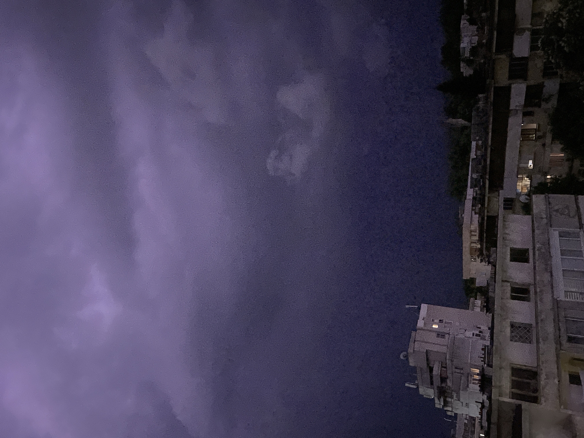 The image depicts a nighttime scene, with a cloudy sky dominating the view, colored in shades of purple and grey. On the right side of the frame, partially visible buildings stand silhouetted against the dark sky, with a few lit windows suggesting occupancy. The scene appears to be in an urban area.