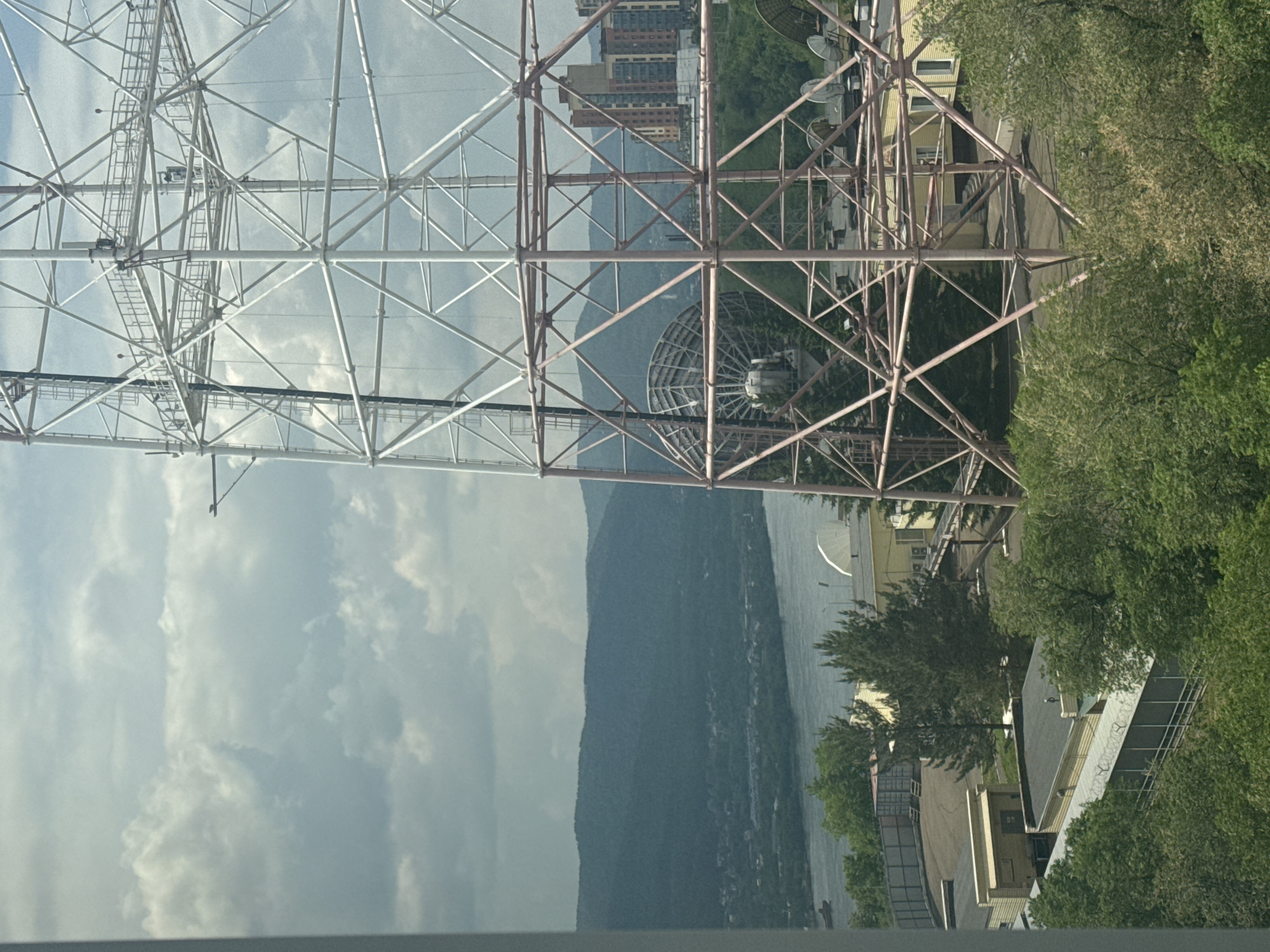 The image shows a large lattice radio tower in the foreground, with a vast, green landscape and a river in the background. The tower is made of white and pink metal struts and has several platforms and antennas attached. In the middle ground, there are several buildings, some with yellow walls, and trees. Beyond the buildings, a wide river flows, with forested hills on the far bank. The sky is cloudy, with patches of blue. The scene is likely taken from a high vantage point, possibly from an apartment building or another tall structure.
Based on visual cues and typical landmarks associated with such structures in Krasnoyarsk, this appears to be the Krasnoyarsk Radio and Television Transmission Tower. This tower is a prominent landmark in Krasnoyarsk, Russia, and is located on a hill overlooking the city and the Yenisei River. The presence of the river, the surrounding hills, and the type of industrial/communication infrastructure are all consistent with this location.