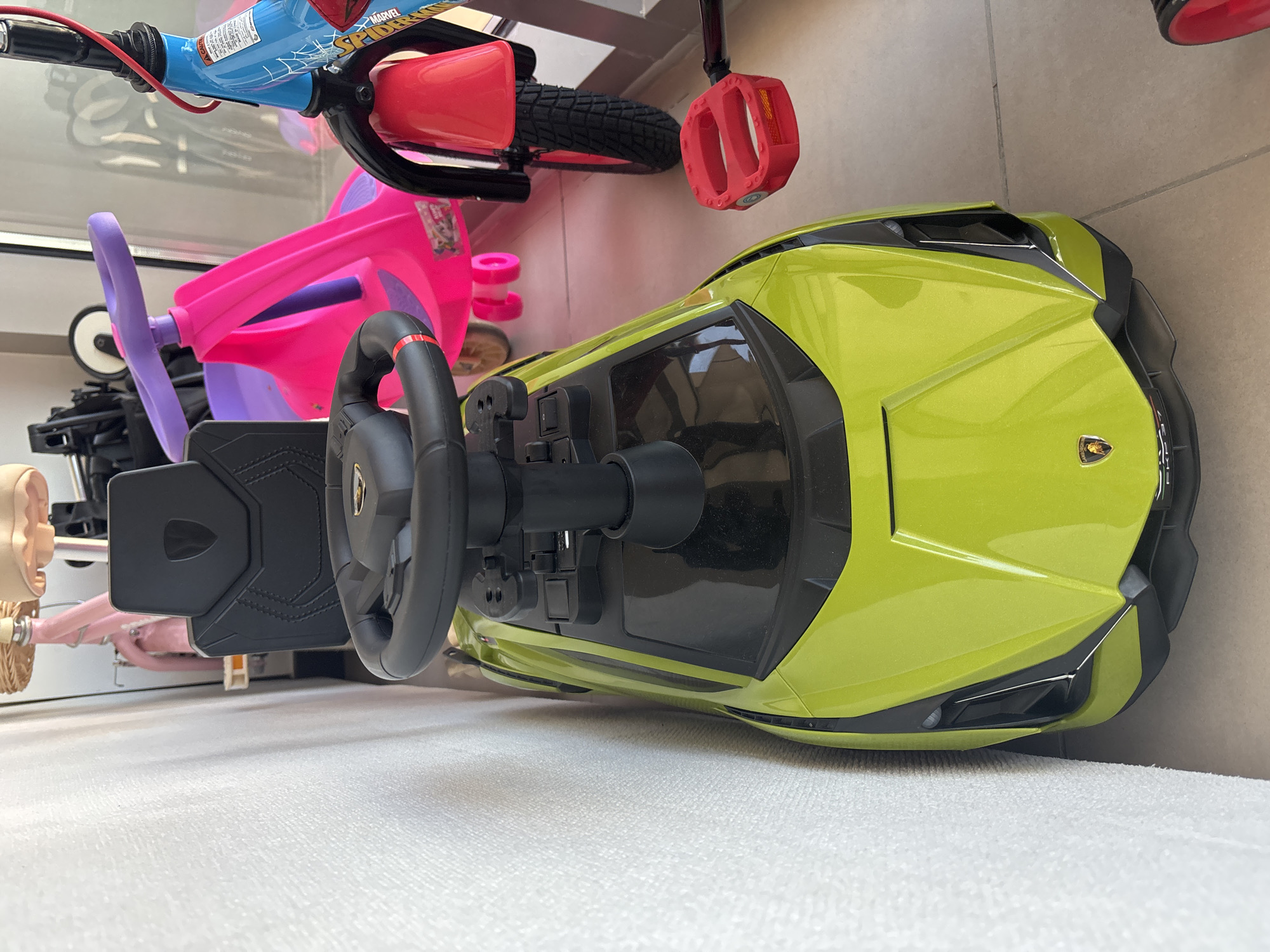 This image depicts a close-up, eye-level view of a bright green toy car with a black seat and steering wheel, positioned against a white textured wall. The car has a Lamborghini emblem on its front. In the background, other children's toys are visible, including a pink ride-on toy with a purple steering wheel and a blue bicycle with a Spiderman theme. A red pedal is also present in the foreground. The flooring appears to be tiled. The scene suggests an indoor setting, possibly a home or a storage area for children's toys, and does not contain any elements that specifically place it in Dubai, United Arab Emirates.