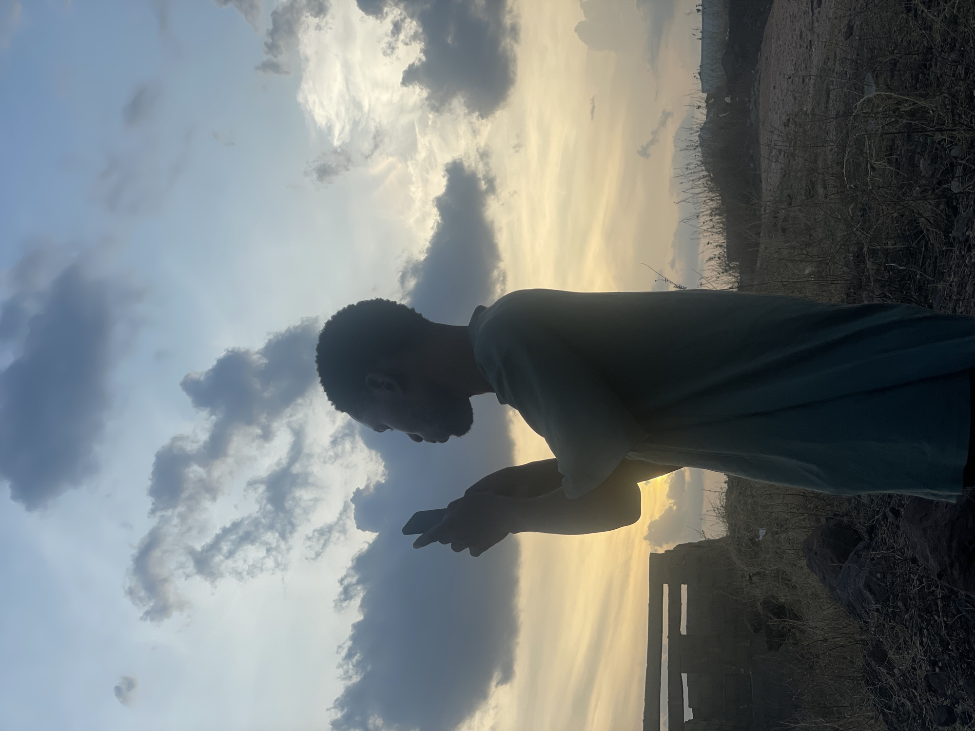 A person is captured in profile, positioned on the left side of a vertically oriented image, silhouetted against a dramatic sky. The individual, appearing male, is holding and looking down at a smartphone with both hands. Their body is largely obscured by shadow, wearing a light-colored garment that also appears dark due to backlighting.
The background is split between a vast sky filling most of the frame and a rugged landscape occupying the bottom right quarter. The sky displays a striking contrast: on the right, a bright, golden-yellow light suggests either sunrise or sunset, while the left features darker, voluminous clouds against a cooler, pale blue sky. The clouds exhibit a range of textures and shades, from soft white to deep grey.
The landscape consists of dry, rocky ground with sparse, dark, spindly vegetation, resembling bushes or tall grasses. Further to the right, dark, rectangular structures, possibly parts of a wall or ruins, are visible against the golden light. The overall scene, captured in what appears to be Lokoja, Nigeria, evokes a sense of quiet contemplation or focused activity amidst a beautiful natural setting at dawn or dusk, with the person engrossed in their device against the grand backdrop. There is no visible text.