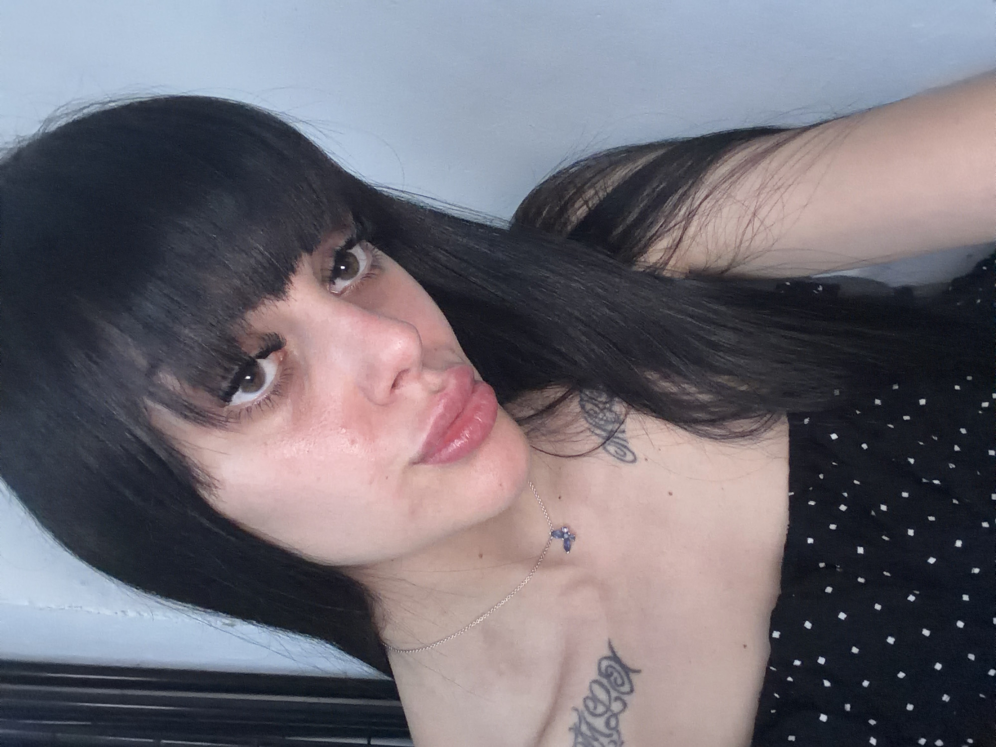 A close-up portrait of a female individual, located in the city of Plovdiv, Bulgaria. The subject has long, straight, dark hair with full bangs covering her forehead. She is looking directly at the camera with her head slightly tilted. Her eyes are dark, and her lips are full and slightly parted. A subtle redness is apparent on her facial skin, particularly around the eyes and cheeks. She wears a black strapless top or dress featuring white square polka dots. A thin silver chain necklace with a small, purple butterfly pendant adorns her neck and chest. Tattoos are visible on both clavicle areas: a script-like design on the left and a partially obscured design on the right. The background consists of a light blue wall to the right and a darker, vertical, possibly textured surface to the left.
