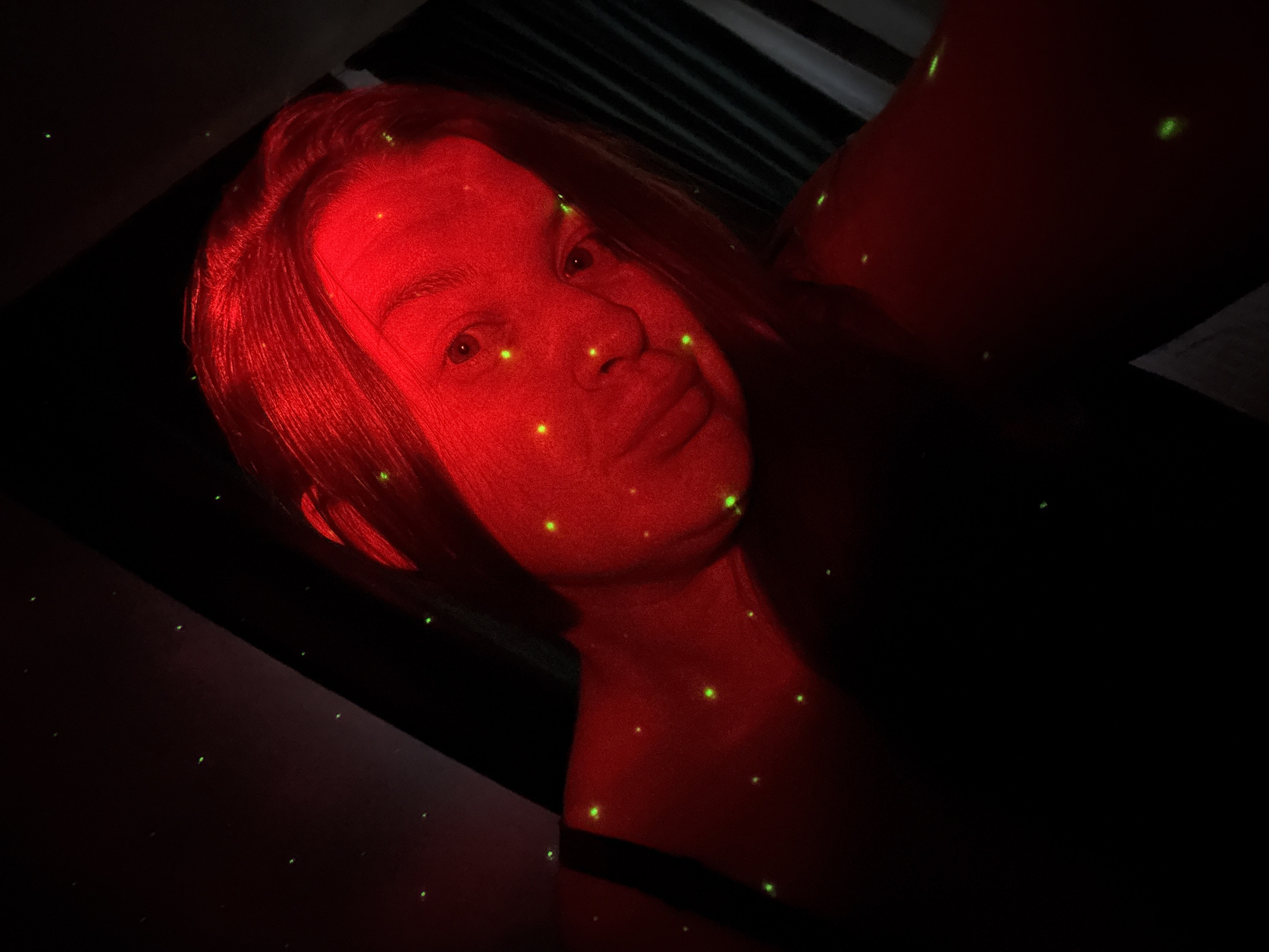 The image shows a person with pale skin under a red light with green dots scattered across their face and body. They are looking slightly upwards. The background is mostly dark. The person appears to be wearing a black strap on their shoulder. The location is not evident from the image and cannot be determined as being in Марковский, Russia.
