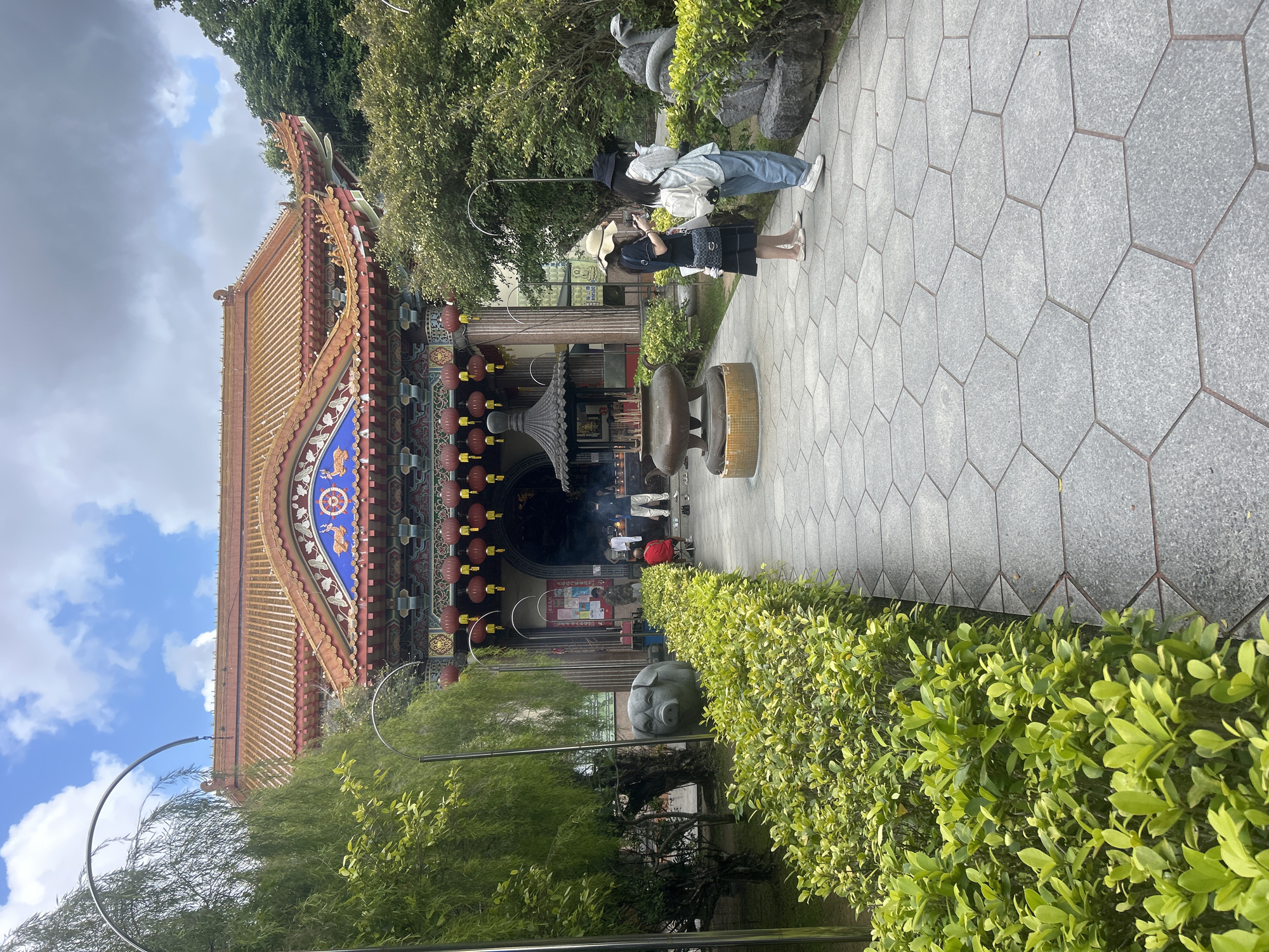 The image shows the exterior of a temple. In the foreground, a pathway made of hexagonal grey tiles leads towards the temple entrance. To the left of the pathway, a neatly trimmed hedge of green bushes runs alongside. Further left, a large stone sculpture of a pig's head is visible behind the hedge, with a weeping willow tree to its left. To the right of the pathway, there are more green bushes and stone sculptures, including one that appears to be a coiled snake.
Two women are standing on the pathway, facing away from the camera and towards the temple. The woman on the left is wearing a black dress and a straw hat, with a shoulder bag. The woman on the right is wearing a light-colored shirt, blue jeans, and a backpack.
The temple itself has a traditional East Asian architectural style with a red tiled roof adorned with decorative elements. Red lanterns are hung along the eaves of the temple's main entrance. Through the entrance, several people are visible, and there is an incense burner in front of the building. The background shows lush green trees and a blue sky with white clouds. The scene appears to be set in a temperate climate.
There is no specific visual evidence within the image to identify the location as George Town, Malaysia. The architectural style is common in East Asian temples and could be found in various locations.