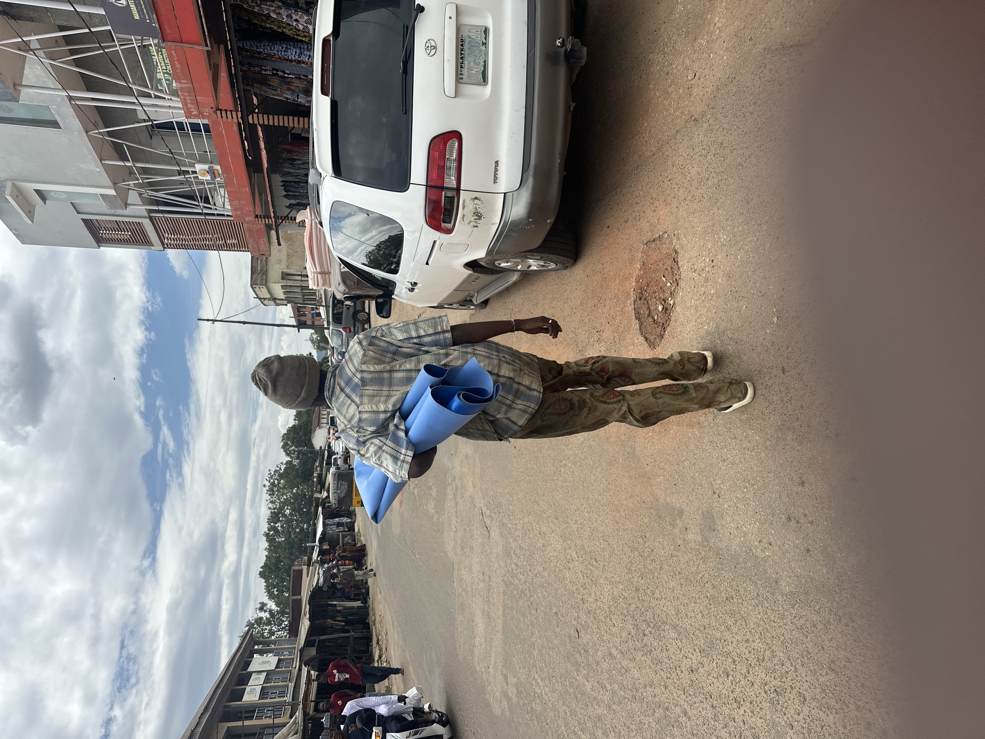 The image depicts an urban street scene in Jos, Plateau State, Nigeria, as indicated by the "PLATEAU" license plate. The primary subject is a male figure viewed from behind, walking away from the camera on the right side of the asphalt road. He wears a grey beanie, a blue and white plaid long-sleeved shirt, brown patterned trousers, and sneakers. He carries a stack of three rolled-up blue items, possibly foam mats or insulation, on his left shoulder.
To the right of the man is a parked white Toyota Sienna minivan with a damaged rear bumper and left taillight area. Its license plate reads "PLATEAU UNG-090AA". A noticeable pothole is visible in the road near the minivan's rear tire.
The street is lined with various buildings. On the left, older, single-story market stalls with corrugated metal roofs feature merchandise hanging outside. Further down this side, a larger, light brown multi-story building is visible. On the right, a more modern, multi-story building with a grey and white facade, red accents, and external white support beams features a sign with partial text "MARIANTE..." and "COLLECTION". Clothes and fabrics are displayed for sale beneath this building.
Several other individuals are visible in the background, walking or standing, including one person on a white scooter on the left. Other vehicles, including a yellow tricycle (keke napep) and cars, are present further down the street. The sky is overcast with scattered clouds. Trees are visible in the distance behind the buildings.