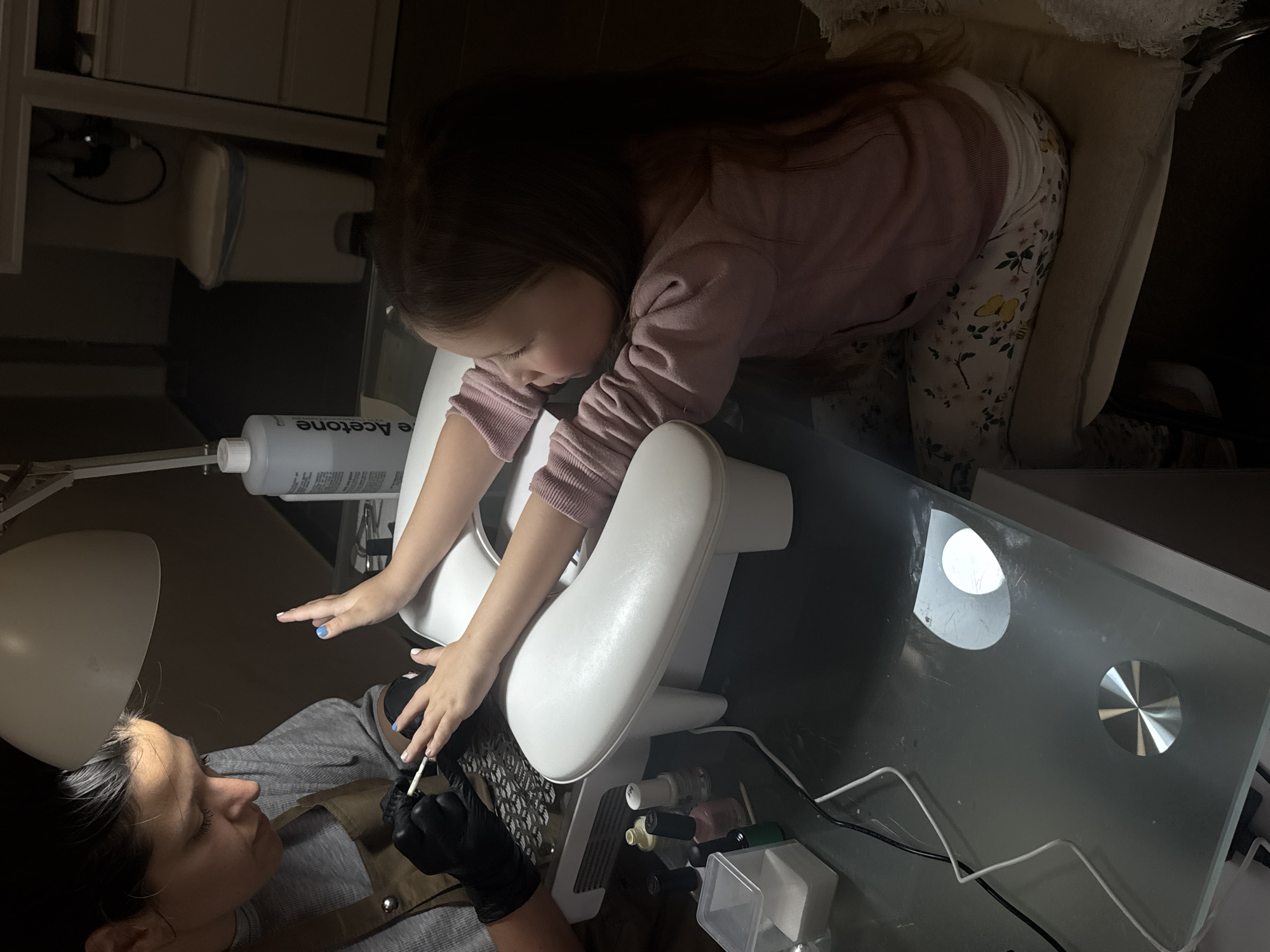 A young girl is receiving a manicure. She is wearing a light pink sweatshirt and floral print pants. She is leaning forward with her hands resting on a white manicure station. A manicurist is applying nail polish to her nails, wearing black gloves. Bottles of nail polish, an acetone bottle, and manicure tools are on the table. The manicure station is a modern design with a glass top and metal accents. The location cannot be determined from the image.
