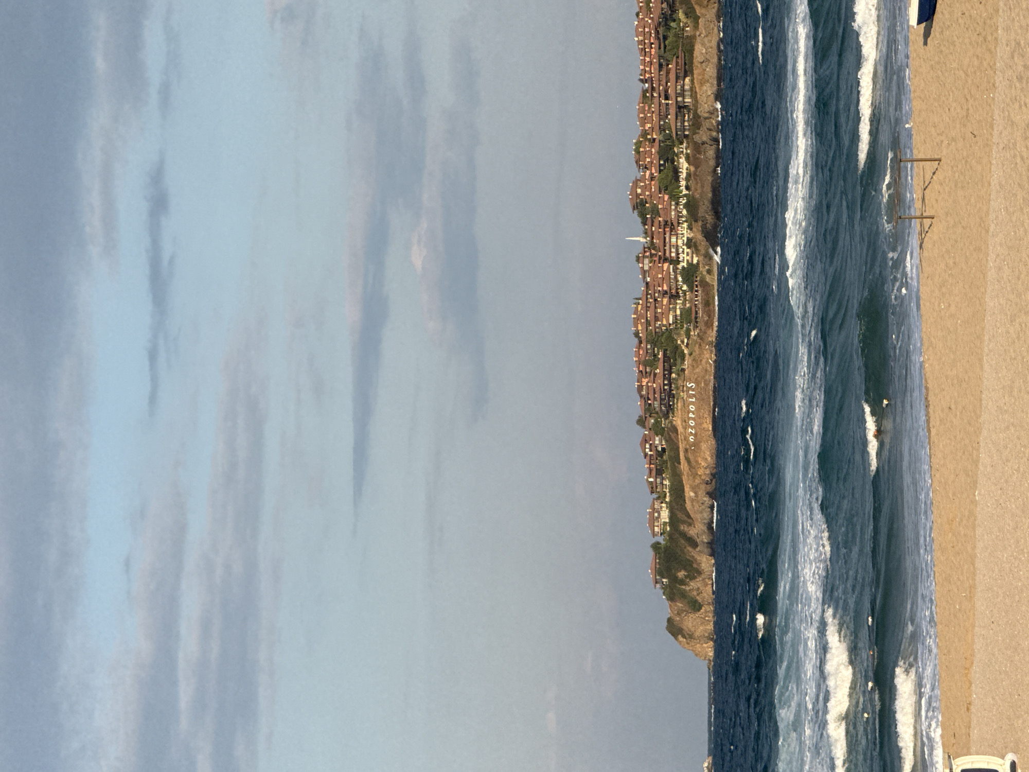 The image shows a coastal scene in Sozopol, Bulgaria. In the foreground, a sandy beach is visible with a small, wooden table-like structure and a small white object. The middle ground features the sea with waves. In the background, a rocky cliff overlooks the sea, topped with a cluster of buildings with terracotta roofs. A sign with the word "SOZOPOLIS" is visible on the cliff. The sky above is partly cloudy.
