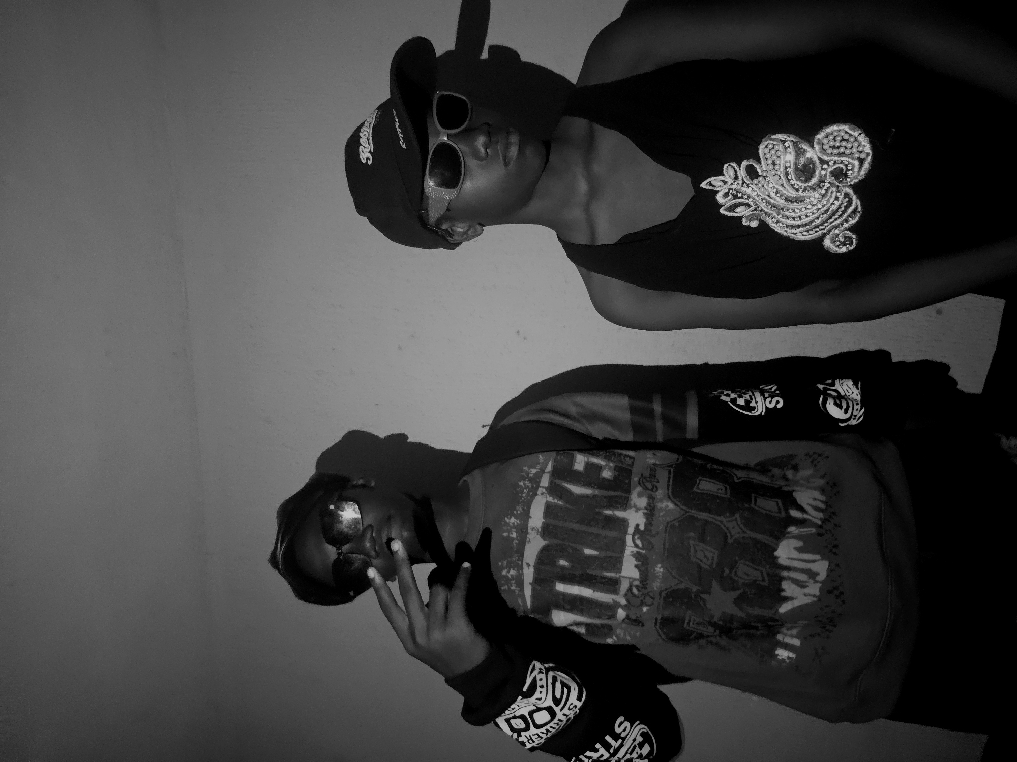 Two people are posing indoors against a light-colored wall in Nkwelle, Nigeria. The first person, located on the upper right side of the frame, is wearing a dark tank top adorned with a decorative beaded design, sunglasses, and a baseball cap. The second person, positioned on the lower right side of the frame, is wearing a long-sleeved shirt with graphic designs on the front and sleeve. They are also wearing sunglasses and gesturing with their hand. The location is indoors with uniform lighting.