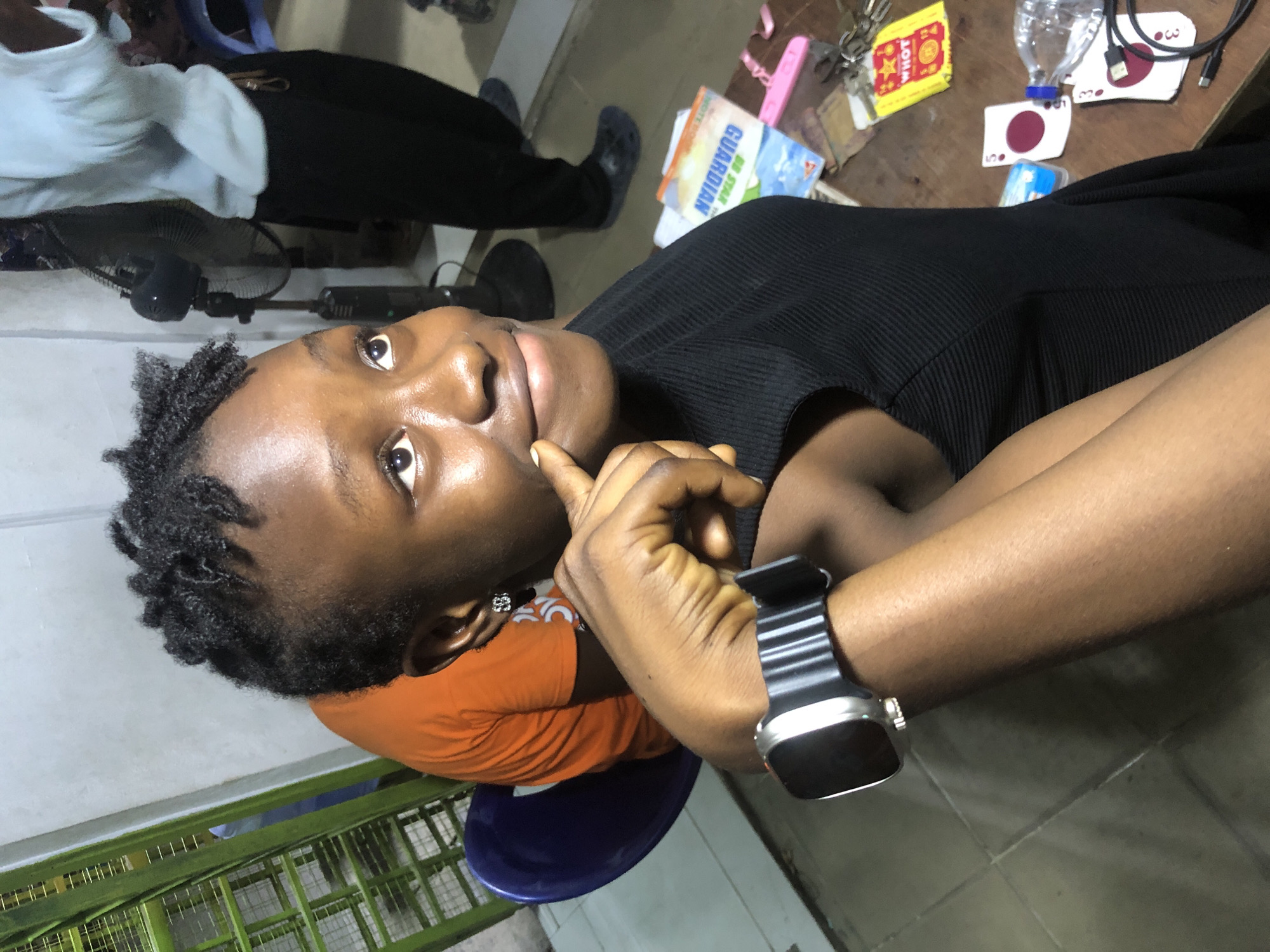 In Ado, Nigeria, an image captures a young Black woman with dark skin and tightly coiled black hair, wearing a black ribbed sleeveless top. She is seated, her left arm resting on a table, and her left hand is raised to her chin, with her index finger touching her cheek. She wears a black digital smartwatch on her left wrist. In the background, a person in an orange t-shirt is partially visible, seated on a blue chair. Further back, another individual in a light blue shirt and dark pants stands near a black standing fan. The environment appears to be indoors, with a tiled floor and a wall that transitions from light grey to white. On the wooden table in the foreground, various items are scattered, including a bottle of water, keys, playing cards with red circles, a yellow packet with the word "WHOT" and stars, and a book titled "GUARDIAN." The lighting suggests an indoor setting with ambient light.