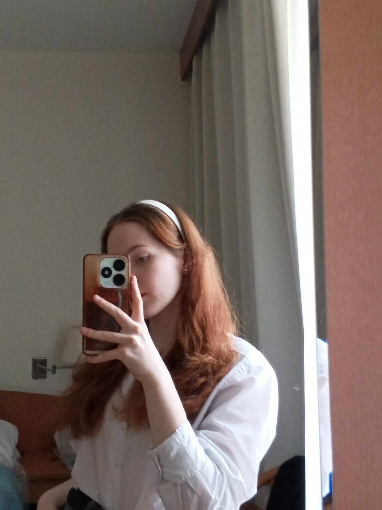 A young woman with red hair and a white headband is taking a selfie in a room. She is wearing a white button-down shirt. The background includes a wall, curtains, a bed, a mirror, and a light fixture. The location in the city of Saint Petersburg, Russia cannot be determined from the visual information.