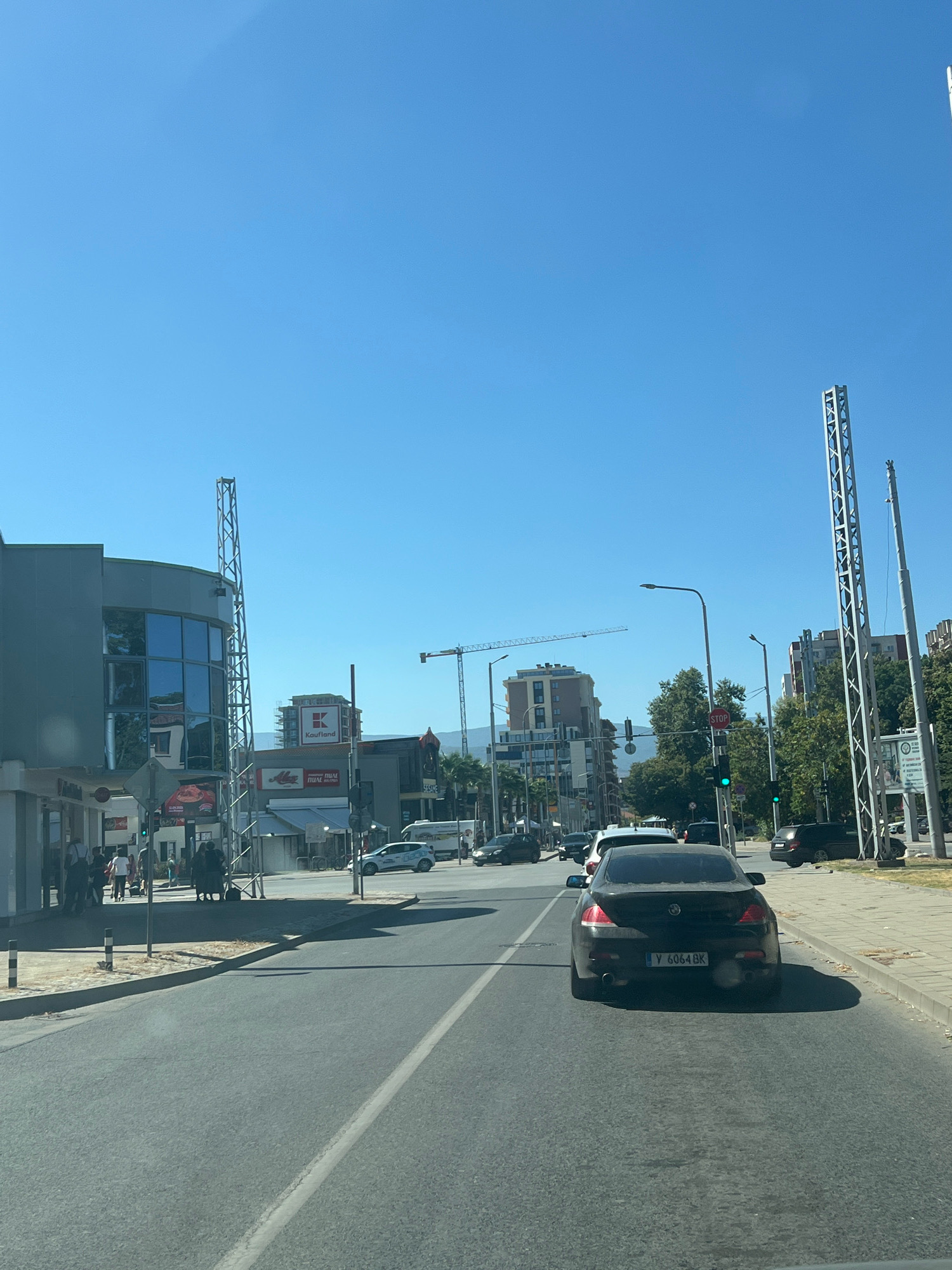 The image depicts a street scene in Plovdiv, Bulgaria, on a clear day. The perspective is from a vehicle moving along a multi-lane road.
In the foreground, the asphalt road takes up a significant portion of the lower half of the image, with a white lane divider visible. A black BMW sedan with license plate Y 6064 BK is in the right lane, slightly ahead and to the right of the viewer's vehicle.
To the left, a modern gray building with large, reflective windows dominates the mid-ground. A tall, metallic truss structure stands in front of this building. Further down the street, several businesses are visible, including one with a prominent "Kaufland" sign and another with "Mega" signage. Pedestrians are seen walking on the sidewalk in front of these establishments.
Across the street, the cityscape features several multi-story buildings, including a tall residential building and other commercial structures. A construction crane is visible in the distance, indicating ongoing development. Traffic lights, with one showing a green light and another displaying a red "STOP" sign, are present. Trees line the right side of the road in the mid-ground and background.
On the right side of the road, a sidewalk and a grassy verge are visible, leading to more buildings and trees. Two tall, slender metallic structures, similar to the one on the left, are positioned along the right side of the street. A white van is parked further down on the right. The sky is a clear, bright blue. The overall impression is a moderately busy urban environment under sunny weather.