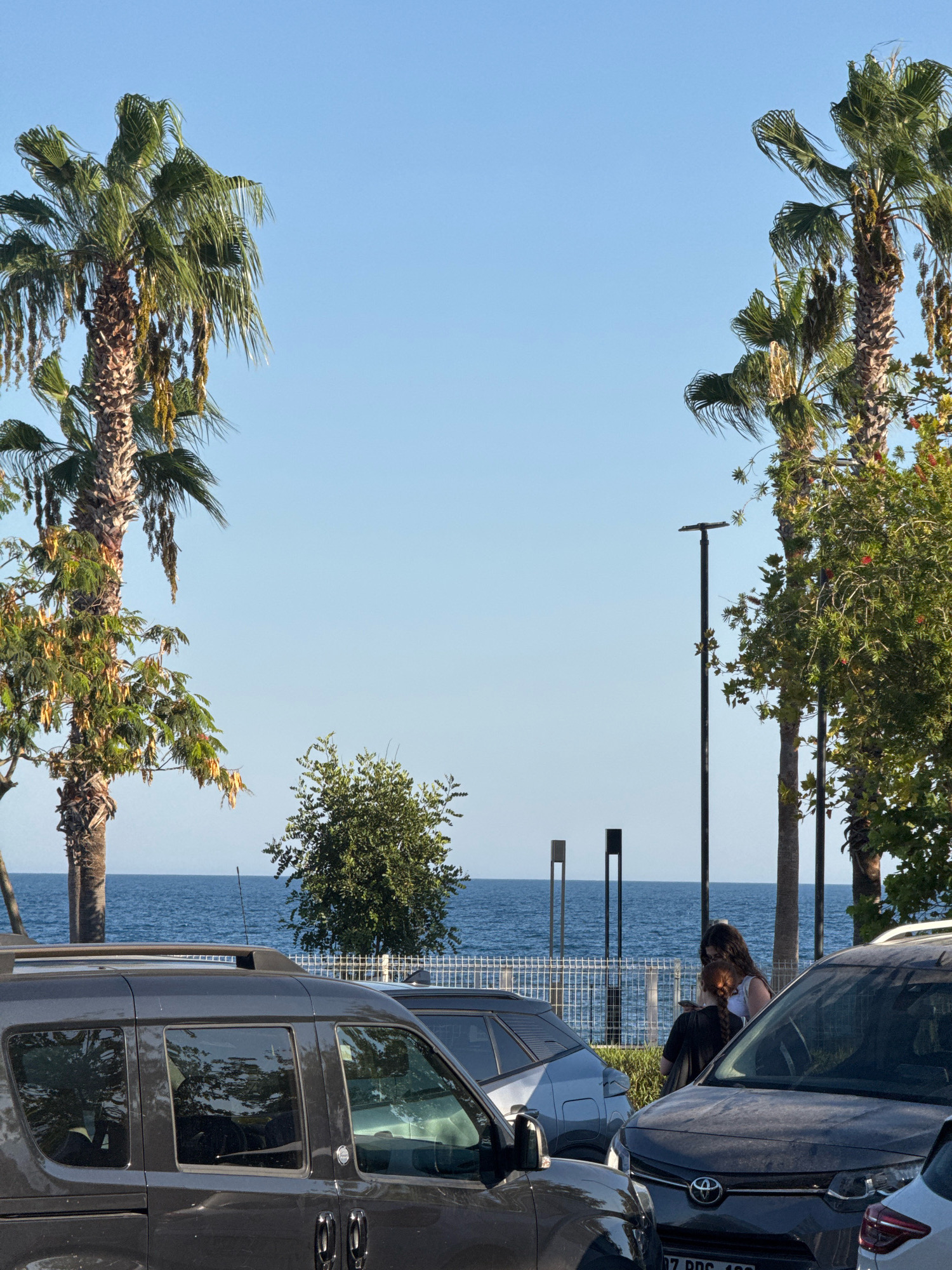 Two women are standing next to parked cars by the sea in Konyaaltı, Turkey. The woman on the left has dark, long hair and is wearing a white tank top and black pants. The woman on the right has red braided hair and is wearing a black jacket. They are both looking down at a phone in the hand of the woman on the left. In the background, the blue sea stretches to the horizon, with a white fence running along the coastline. Palm trees and other greenery are visible, as are streetlights and several parked cars, including a dark grey minivan in the foreground. The sky is clear and blue.