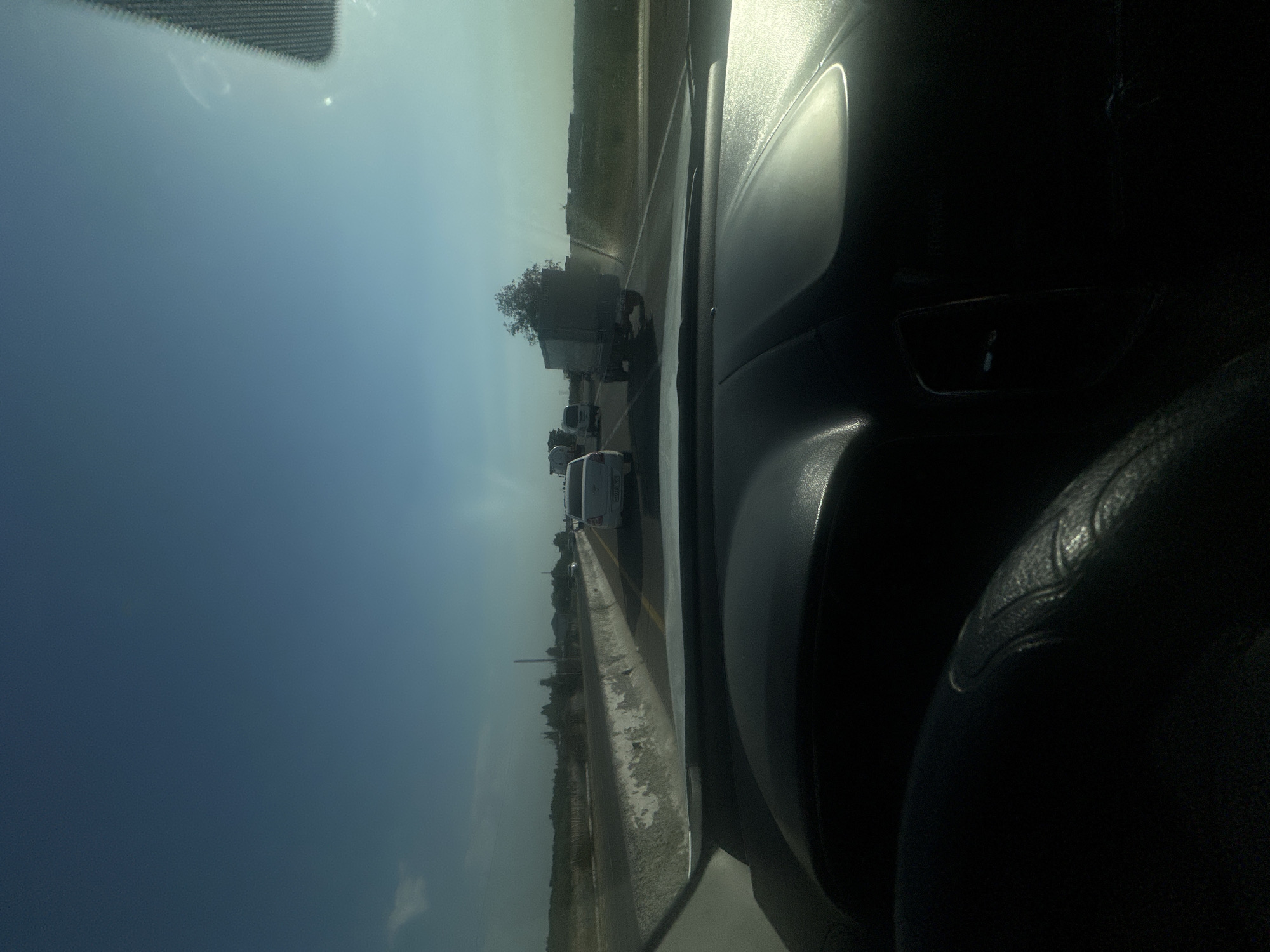 This media file depicts a view from inside a vehicle, driving on a highway. The scene shows a bright, clear day, likely late morning or early afternoon due to the strong sunlight. The sky is a vibrant blue, with some wispy white clouds.
The foreground is dominated by the interior of the car, with a dashboard and steering wheel visible on the right side. The dashboard is dark and appears to have a reflective surface.
The view outside the car's windshield shows a highway with multiple lanes of traffic. Several vehicles are visible, including a large truck with a box trailer carrying what appears to be vegetation. Other cars and trucks are also present on the road, suggesting moderate traffic. A concrete barrier separates the lanes of traffic from the left side of the road. Beyond the highway, there is a flat landscape with low vegetation and some trees.
There are no people clearly visible within the frame. The overall impression is one of travel on a sunny day. No text is legible in the image. The location context provided is Tashtugizak, Uzbekistan.