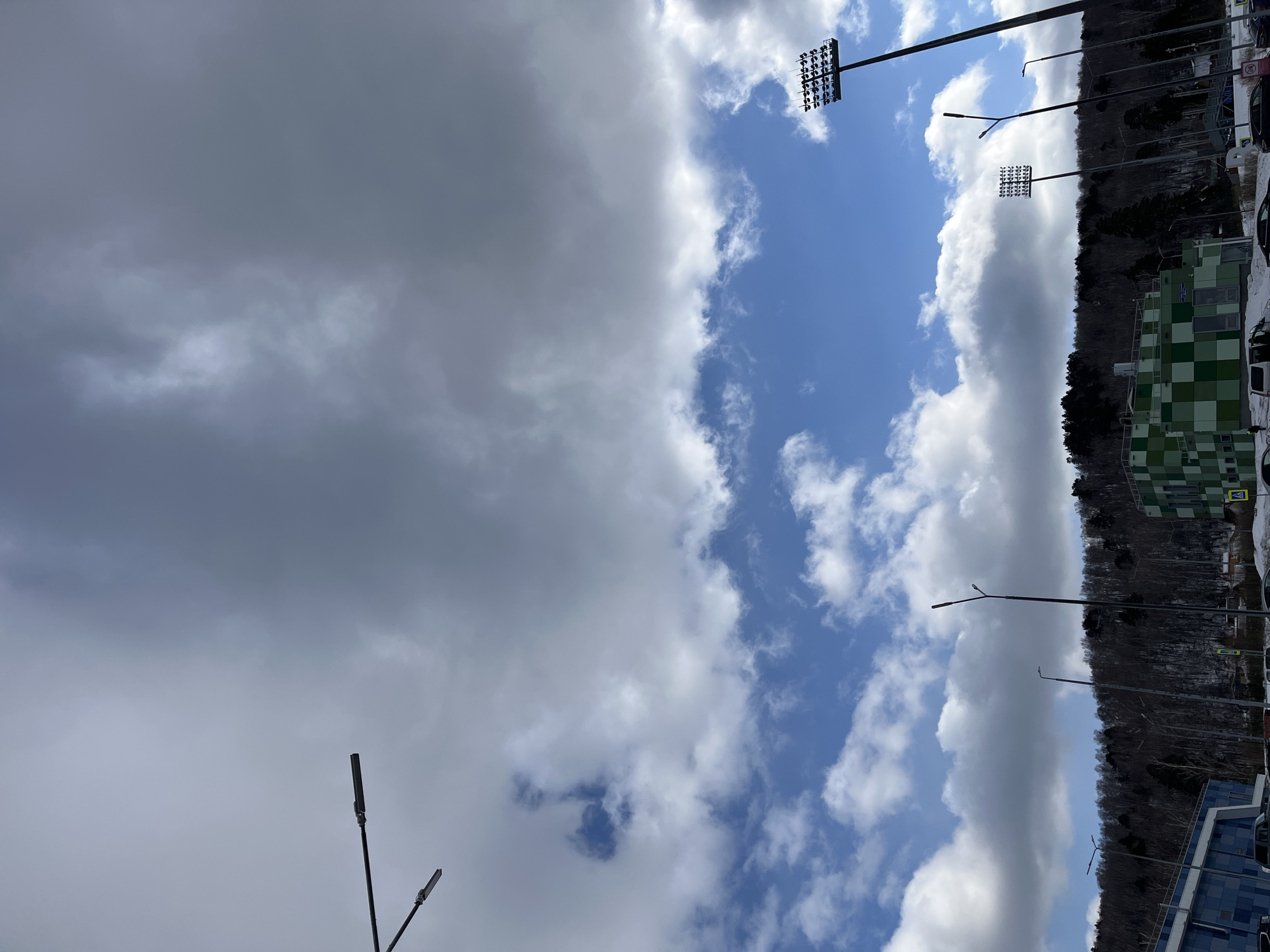 The image depicts a partially cloudy sky above a landscape in Krasnoyarsk, Russia. The sky is bisected; one side shows a dense, grey cloud cover, and the other shows a clear blue sky with scattered white clouds. Several tall light poles with multiple lamps are visible, pointing up towards the sky. A forested hillside borders the scene, next to a checkered green and white building with visible windows and signage. Adjacent to the building is a parking lot with cars. Another building with blue reflective windows is partially visible at the bottom of the frame.