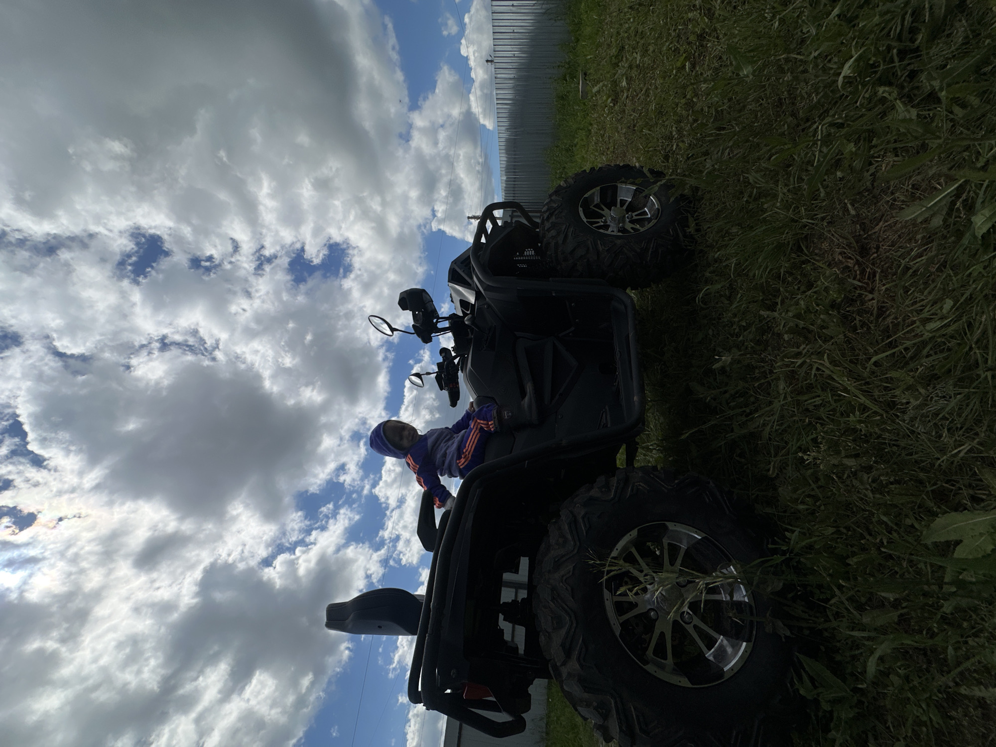 A young child, dressed in a blue hooded sweatshirt and striped pants, is seated on a black all-terrain vehicle (ATV). The ATV is parked on a grassy area with a metal fence in the background. The sky above is partly cloudy, with the sun shining through in some areas. The scene appears to be an outdoor recreational space. The location of Kharnavy, Russia, is not discernible from the visual information provided in the image.