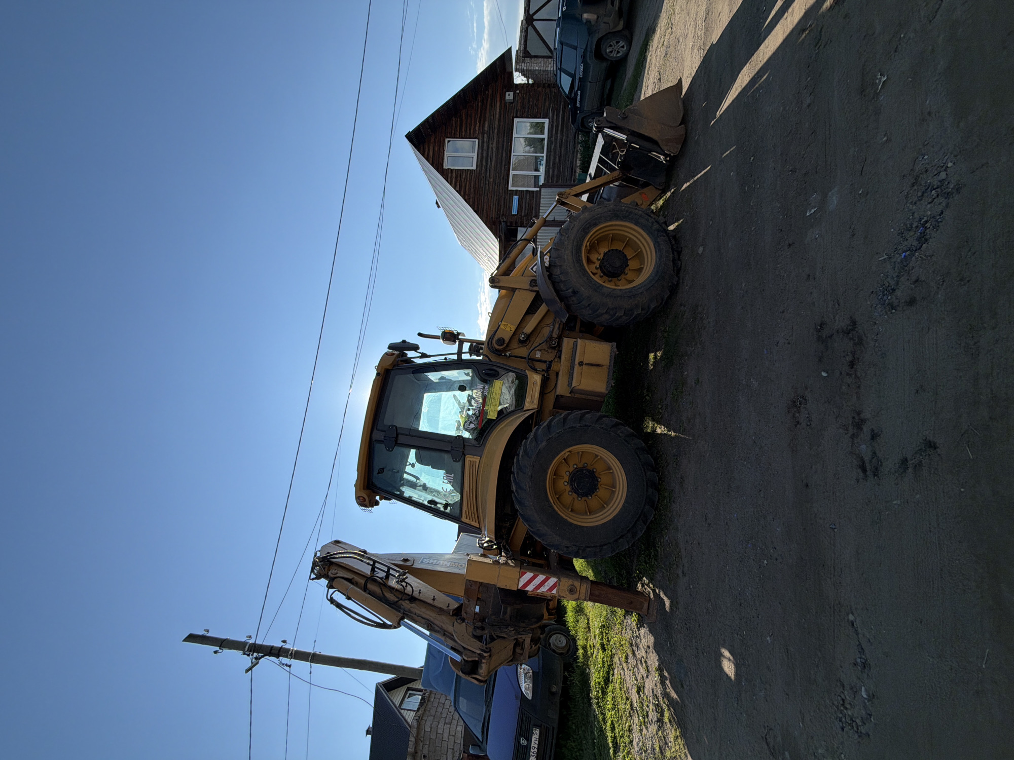 The image, oriented incorrectly and showing a clear, sunny day, features a yellow backhoe loader parked on a dirt and gravel path. The backhoe has large tires, a front loader bucket, and a rear excavator arm with a red and white striped warning decal on its stabilizer. The sun, high in the sky, casts long shadows from the backhoe and nearby structures.
In the background, a two-story wooden house with white-framed windows and a steep gable roof is visible. A dark-colored vehicle, possibly a van or SUV, with "СЛУЖБА" (SLUZHBA, meaning "Service") written on its side, is parked near the house. Partially obscured by the backhoe on the left side of the frame, a blue car displays a Russian license plate, "369 YH 59". Utility poles and power lines are prominent against the bright blue sky. The ground consists of uneven dirt and gravel, with some green grass. No people are present in the scene. The setting is Chaikovsky, Russia.