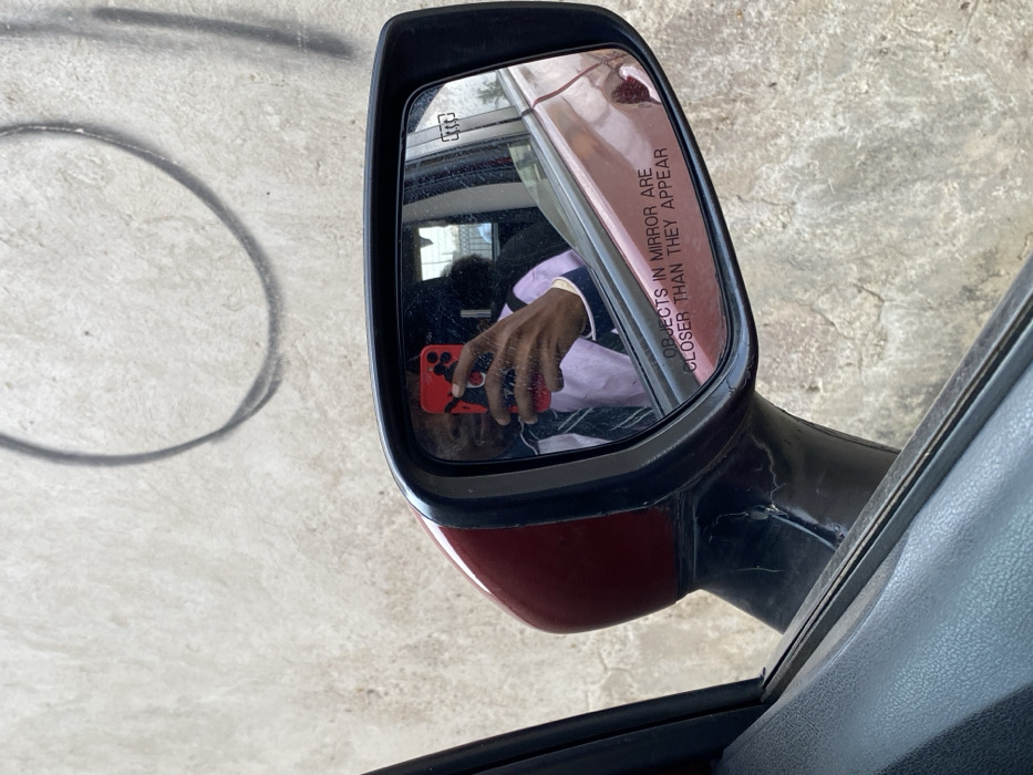 The image displays a close-up view of a dark red vehicle's passenger-side mirror, exhibiting scuffs and wear on its casing and reflective surface. The mirror glass contains the etched warning "OBJECTS IN MIRROR ARE CLOSER THAN THEY APPEAR" and a heating element icon. Reflected within the mirror is a person, positioned inside the vehicle, wearing a light-colored dress shirt and a dark tie. This individual is actively holding a red smartphone, featuring a black-and-white patterned case, angled towards the mirror to capture their reflection. The dark-colored interior door panel of the vehicle is visible in the immediate foreground. The background outside the vehicle consists of a textured, light-toned wall, exhibiting stains and a prominent, incomplete black spray-painted circular mark. The scene is located in Unknown, Ghana.