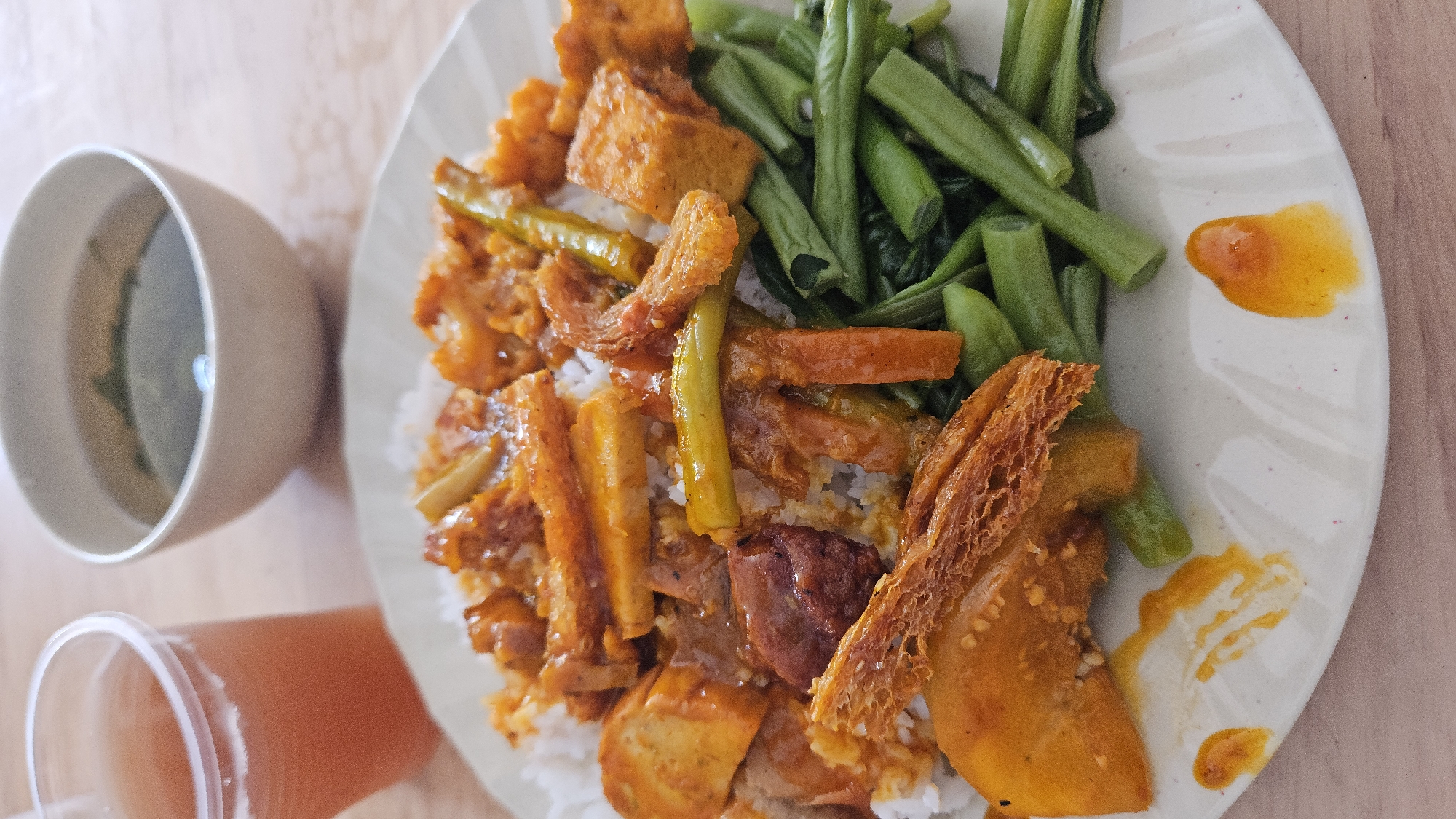 A plate of Vietnamese cơm tấm (broken rice) is presented alongside a cup of soup and a drink. The cơm tấm dish is topped with various fried tofu, beans, and vegetables, covered with a sauce. It is placed on a light-colored wooden surface in a Cam Ranh Ward, Vietnam setting.