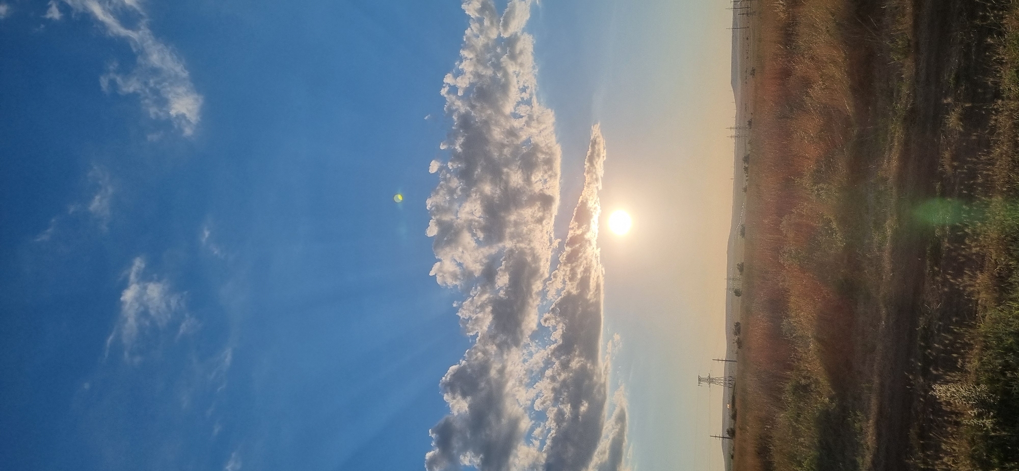 This is a vertical, wide-angle photograph of a rural landscape under a partly cloudy blue sky at sunset. The sun, a bright, glowing orb, is positioned slightly left of center in the middle of the frame, partially obscured by a horizontal band of fluffy white and grey clouds. Beams of light radiate from the sun, creating a dramatic sunburst effect in the sky. In the foreground, a field of dry, golden-brown grass stretches across the bottom two-thirds of the image. The terrain is slightly hilly, with low vegetation and several tall, latticed electrical pylons visible in the middle distance, suggesting the presence of a power grid. The horizon line is low, emphasizing the expansive sky. A faint green lens flare is present in the lower right corner. The location is identified as Blizhnee, Ukraine. There are no people or animals visible in the scene.