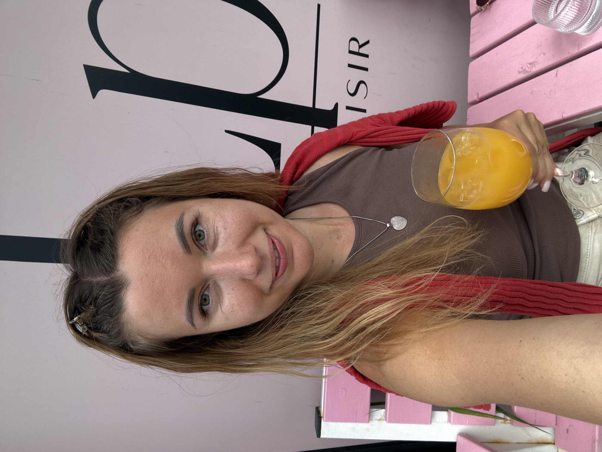 A woman with blonde hair and blue eyes takes a selfie outdoors, holding a glass of orange juice. She is wearing a brown tank top and a red cardigan draped over her shoulders. A silver heart-shaped pendant necklace is visible. The background features a pink wall with a black logo that reads "L ISIR". A pink wooden bench is also present in the scene. The lighting is natural, suggesting it is daytime.