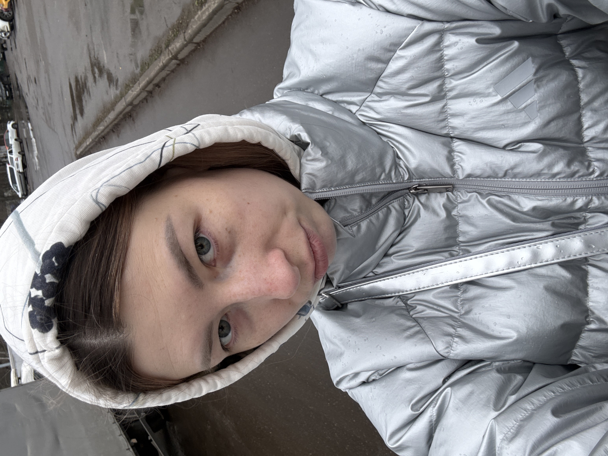 A pale-skinned young woman with brown hair and gray-green eyes looks directly at the camera. She wears a light-colored hooded sweatshirt with dark floral patterns around the opening. Over the sweatshirt, she has on a shiny silver puffer jacket with visible water droplets. She is outdoors, near a street with a sidewalk and parked cars visible in the background. The setting suggests an urban area, possibly in a Russian city. The photo appears to be taken during or after a period of light rain.