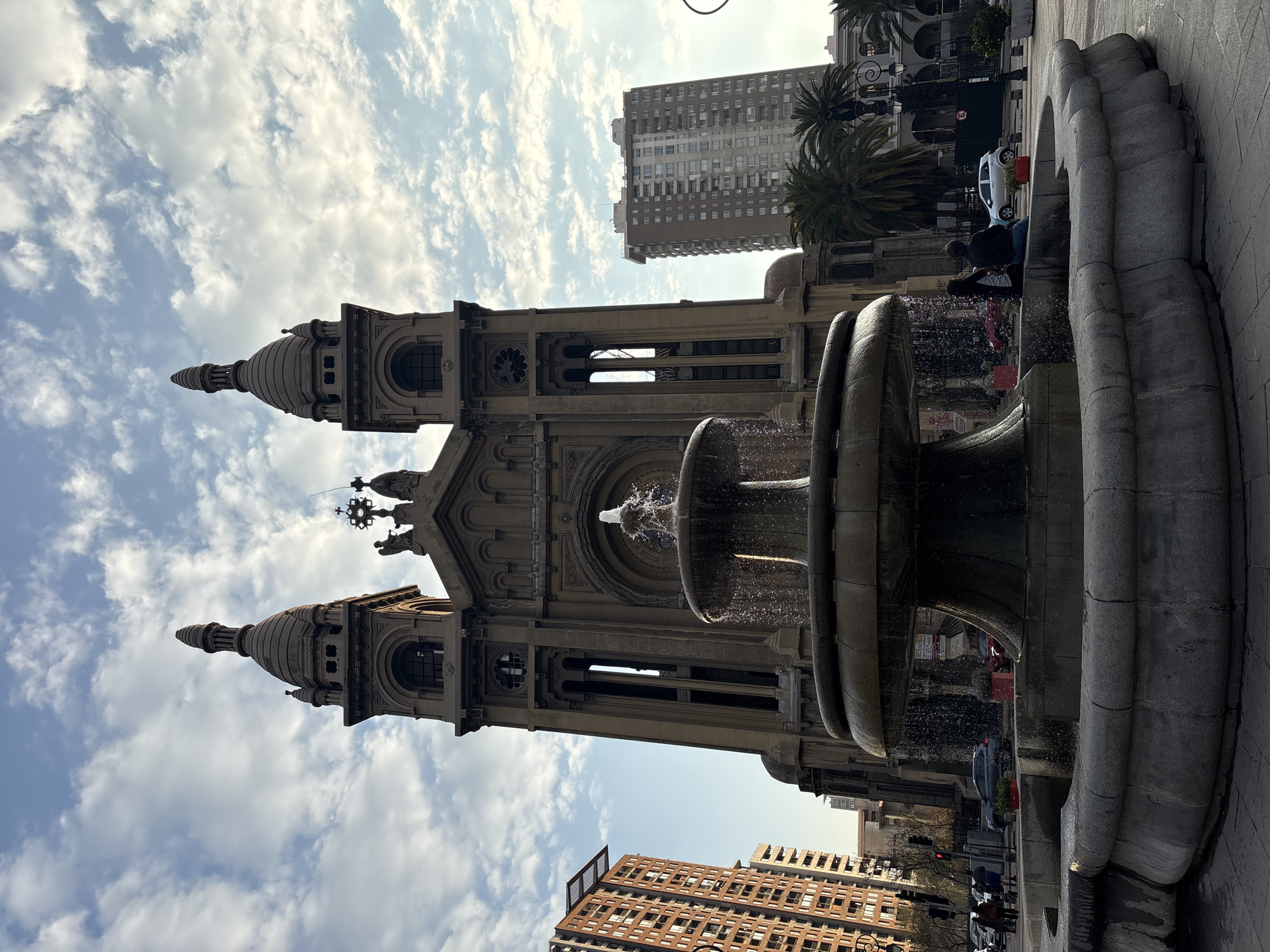 The image shows a large, ornate fountain in the foreground, with a grand stone church in the background. The church features twin bell towers and intricate architectural details. To the left of the church, a modern, multi-story building with a reddish-brown facade stands tall. To the right of the church, another tall, contemporary building with a beige facade is visible, along with palm trees. The sky is partly cloudy with scattered white clouds against a pale blue background. Several cars are present on the street behind the fountain, and two people are seated on the edge of the fountain, facing away from the camera. The fountain is actively spraying water, creating a shimmering effect. The scene appears to be a public square in Santiago, Chile, given the architectural style and the presence of palm trees.