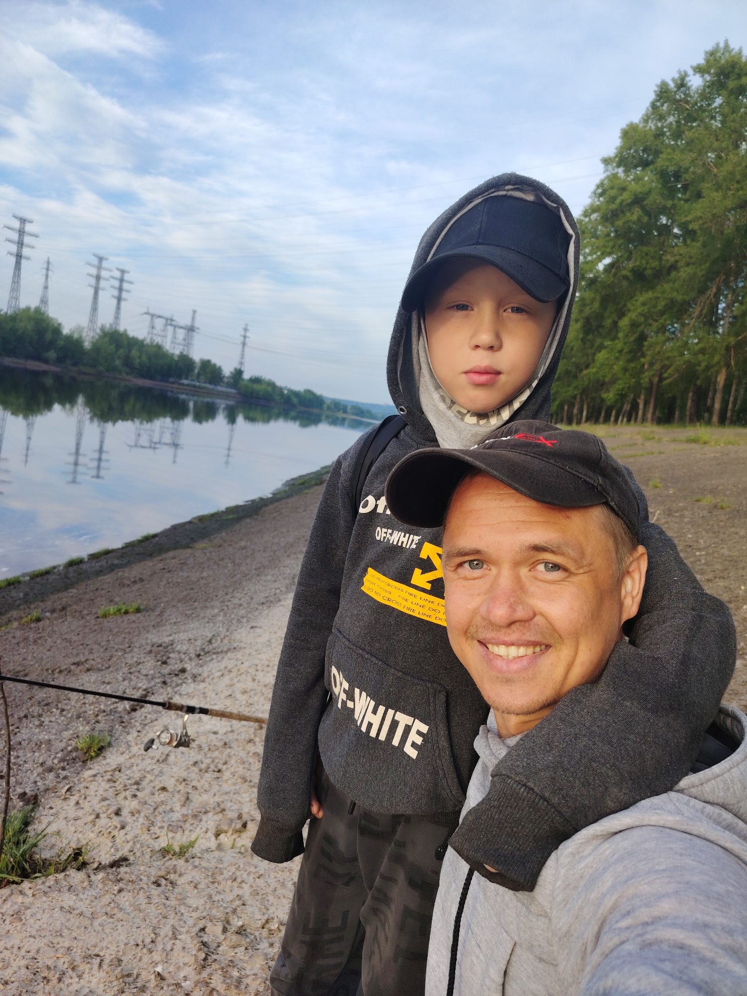An outdoor, eye-level selfie captures an adult male and a boy standing on a riverbank, likely in Chaikovsky, Russia. The adult, positioned in the foreground, smiles broadly at the camera, wearing a dark cap and a grey hooded sweatshirt. His left arm is around the boy's shoulders. The boy, who appears younger, stands slightly behind and to the left of the man, looking towards the camera with a neutral expression. He wears a dark cap under a grey hooded sweatshirt with the hood up.
The background reveals a tranquil river with reflections of the sky and distant trees. On the far bank, several large power line towers are visible, suggesting an industrial or semi-rural setting. The sky above is partly cloudy with soft, diffused light, indicating either an overcast day, morning, or late afternoon. To the left of the boy, a fishing rod is visible, resting on the ground with its reel, hinting at a fishing activity. The ground beneath them is a mix of dirt and gravel.
Text visible on the boy's sweatshirt includes the brand "OFF-WHITE" and repeated phrases like "DO NOT CROSS FIRE LINE DO" in a yellow block font. The overall mood of the image is calm and familial.