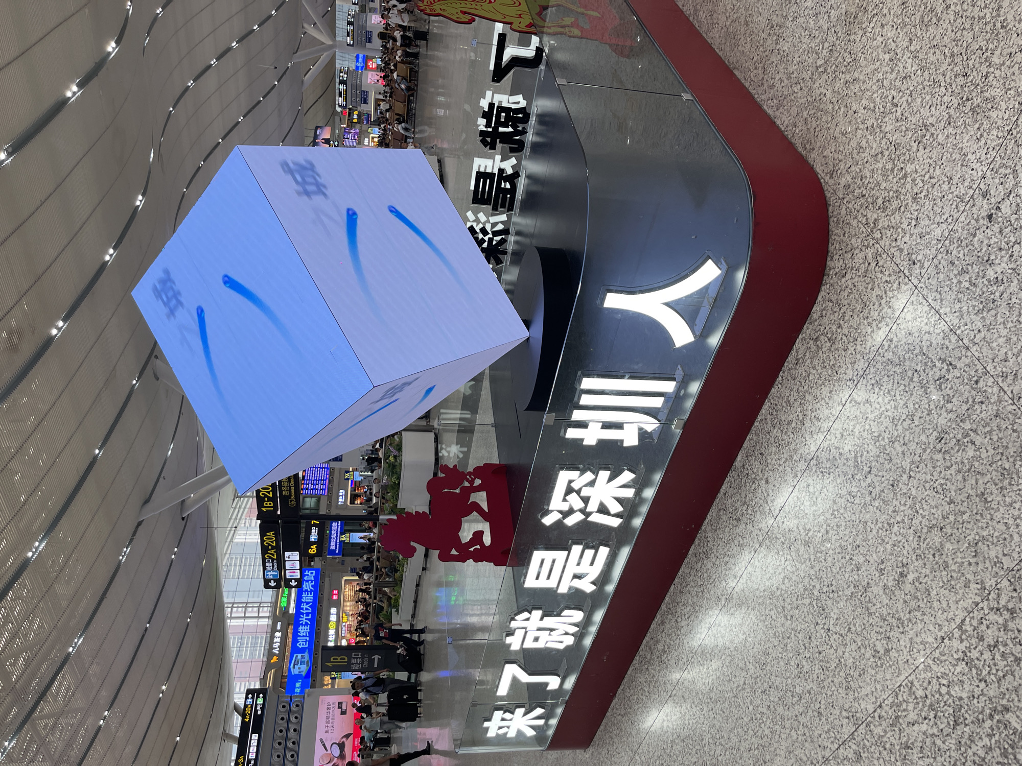 This media file depicts a busy interior scene, likely an airport or train station in Longhua District, China. The primary subjects are people milling about, interspersed with various objects and signage. A large, illuminated cube displaying blue abstract shapes is a central visual element. Adjacent to it, a prominent display features bold white Chinese characters on a black background, which read "深圳北站欢迎您" (Shenzhen North Station Welcomes You) and "来了就是深圳人" (You are a Shenzhen person once you are here). Red signage, possibly indicating departure gates or information points, is also visible.
The setting is a modern, well-lit indoor space with a high, curved ceiling featuring numerous lights. The floor is a speckled gray tile. Several directional signs with Chinese text and arrows guide people, indicating check-in areas, platforms (e.g., "2A-20A", "6A"), and commercial establishments like "全家" (FamilyMart) and "家仕美超市" (Jia Shi Mei Supermarket).
The activity appears to be the normal flow of travelers, with some people standing, others walking, and some seated in waiting areas. The overall atmosphere seems bustling and functional. The time of day is not explicitly stated, but the bright indoor lighting suggests it is daytime. No specific emotions are evident, but the scene conveys a sense of transit and a welcoming message to visitors. The presence of signs in both Chinese and English, along with brand names, points to a modern and international hub.