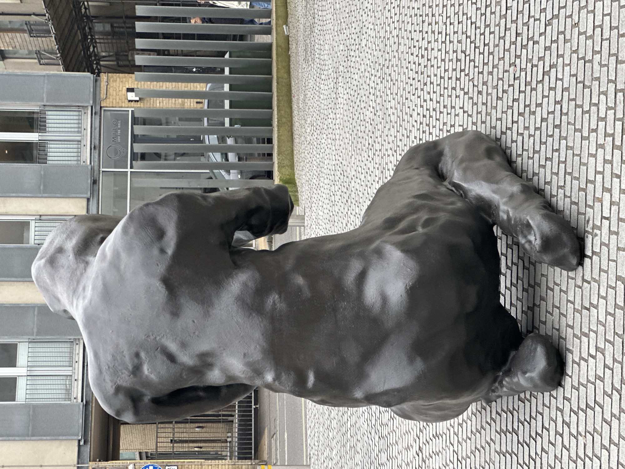 A dark, metallic sculpture of a crouching human figure, viewed from behind, is centrally positioned on a light grey, rectangular sett pavement. To the left, an asphalt street with a white line and a yellow pole is visible. In the mid-ground, a building with a light-colored brick base and a grey upper facade features multiple windows with metal grilles. The ground floor displays a shopfront with the 'MILO and THE BULL' logo, identifying the location as 23 St Thomas Street, London SE1 9GT, United Kingdom. A dark grey vertical slat fence runs in front of the building, partially obscuring a dark grey sedan and a green utility bin. Two indistinct individuals are observed walking in the far right background, behind the fence. The scene is under overcast lighting conditions.