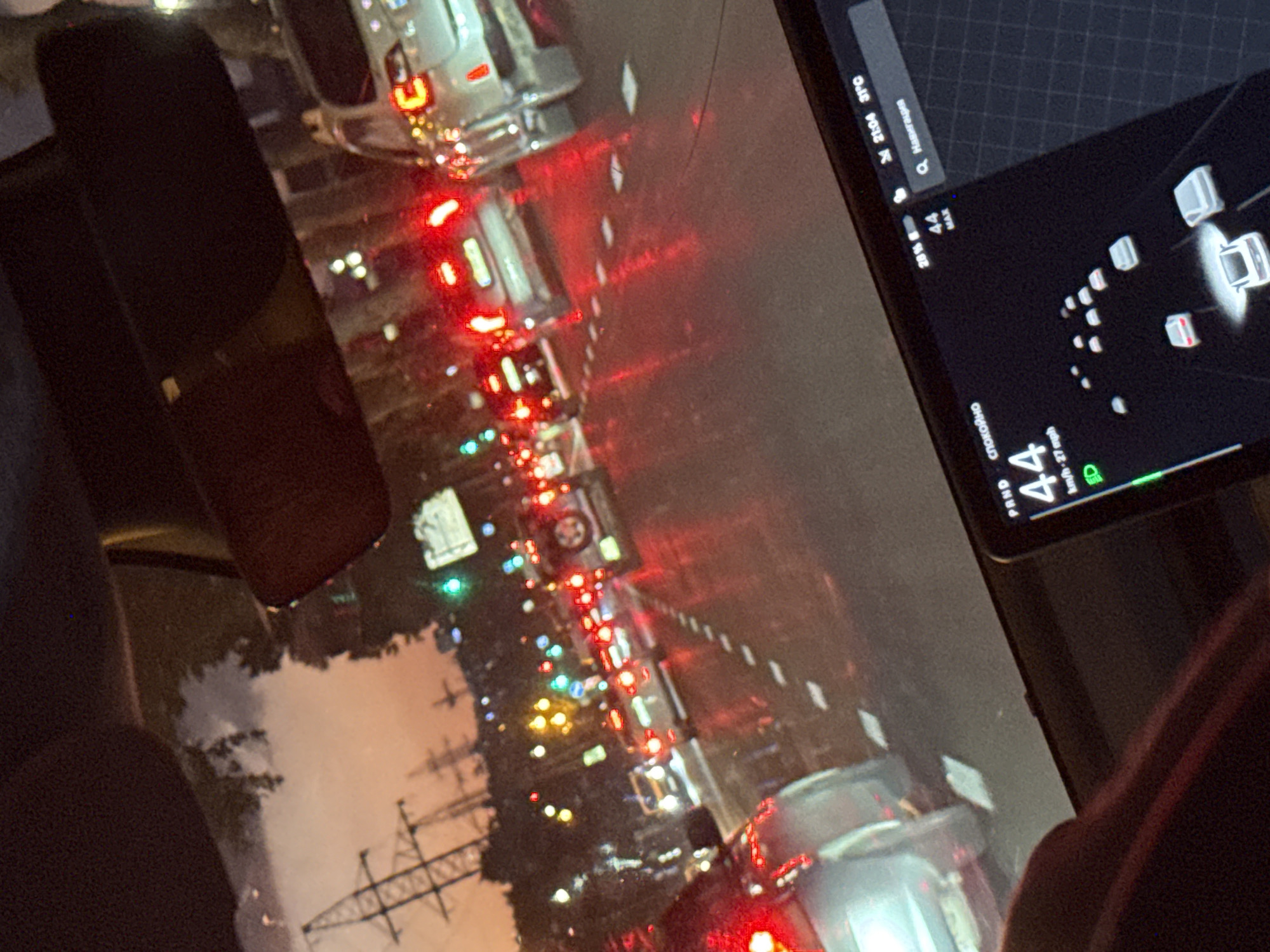 The image shows a view from inside a car, looking towards the dashboard and out the windshield. The dashboard features a screen displaying navigation information, including the time "21:04", temperature "31°C", and a map with car icons. The car appears to be in traffic, as indicated by the brake lights of other vehicles visible on the road ahead. The road is wet, suggesting it may have recently rained or is currently raining. The overall scene is dark, consistent with nighttime, and the artificial lights from the vehicles and the dashboard create a somewhat surreal ambiance. There are also power lines visible in the distance, hinting at an urban or suburban setting. The text on the dashboard screen includes "PAND", "спокойно" (which means "calm" or "quiet" in Russian), "MAX", and "km/h-27 mph", as well as "Q. Навигация" (which means "Navigation" in Russian). The presence of Russian text suggests the car's system may be set to that language, which is not uncommon in Tbilisi, Georgia, due to historical ties. The driver's hand or arm is partially visible on the right side of the frame, suggesting someone is operating the vehicle. The scene evokes a sense of being stuck in traffic on a damp evening.