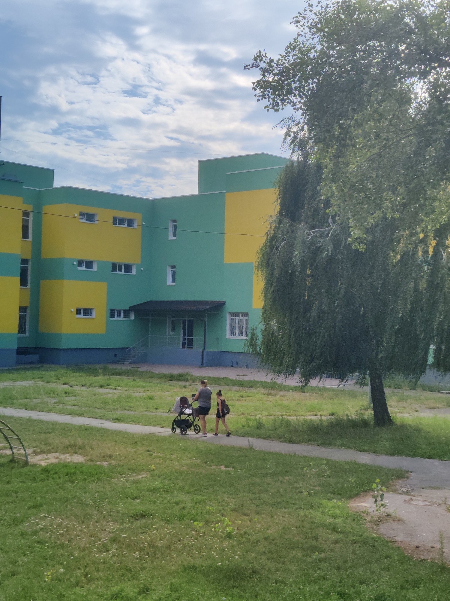 The image shows a modern building painted in alternating bands of green and yellow, likely a school or administrative building, under a partly cloudy sky. In the foreground, a paved walkway cuts through a grassy area. On the walkway, a woman is pushing a stroller, accompanied by a young girl walking beside her. A large tree with drooping branches is prominent to the right of the building. The scene appears to be in an urban or semi-urban setting in Kyiv, Ukraine, given the architectural style and common urban flora.