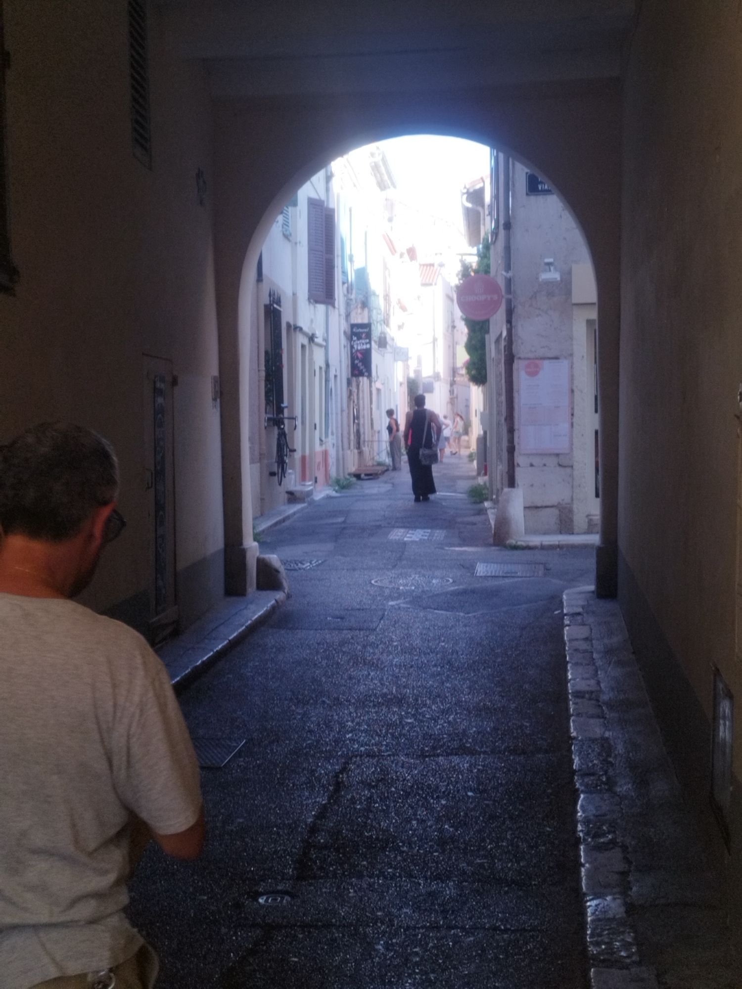 The image shows a narrow cobblestone alleyway in Antibes, France, with buildings on either side and an arched opening at the end. The alley is lined with shops and restaurants. The ground is wet, suggesting it has recently rained. In the foreground, a man with short, dark hair is seen from the back, wearing a light-colored t-shirt. Further down the alley, a woman in a dark dress walks away from the camera, carrying a dark bag over her shoulder. Another woman in a light-colored dress is visible further ahead. A bicycle is attached to the wall of one of the buildings, and a red sign with "CHOOFT'S" written on it hangs above a shop entrance. The overall impression is one of a bustling street scene in a European city.