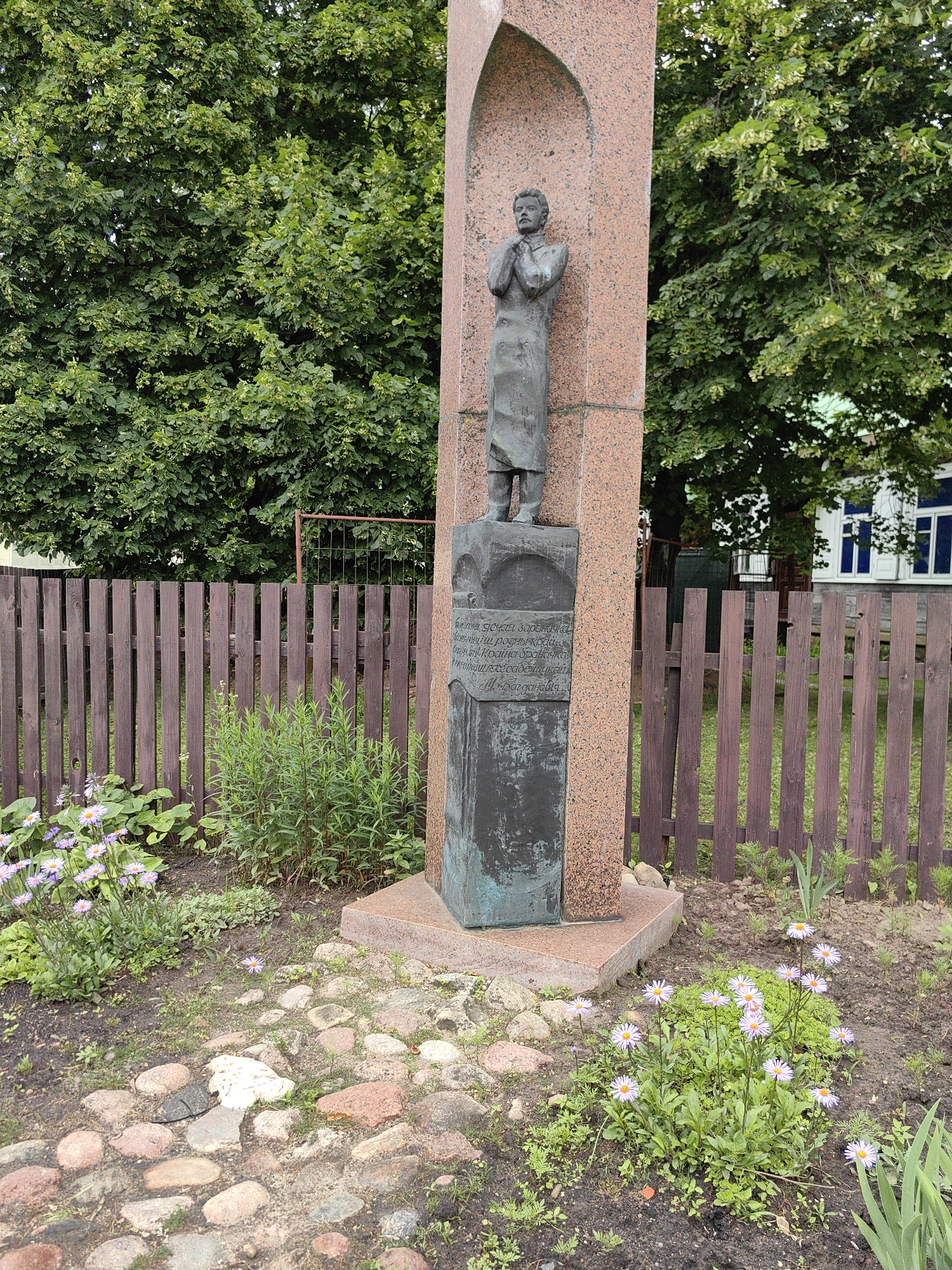 The image shows a statue of a man in prayer, set against a backdrop of lush green trees and a wooden fence. The statue is positioned in front of a large, reddish-pink stone monument with an arched alcove. Below the statue, on a dark bronze plaque, there is an inscription in Cyrillic script. The ground is covered with small stones and some blooming purple and white flowers. In the background, a building with white-trimmed windows can be seen behind the fence. This monument is located in Minsk, Belarus, at the intersection of Independence Avenue and Komsomolskaya Street.