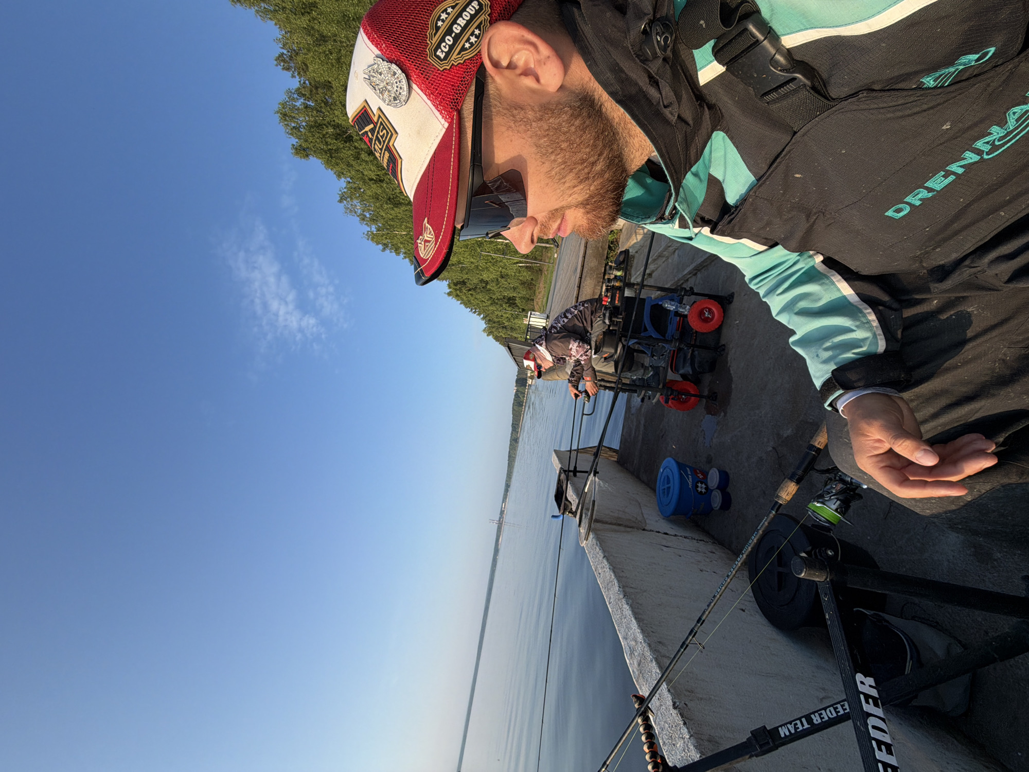 The image depicts a bright, sunny day with a clear blue sky. The scene is set beside a body of water, likely a river or lake, with a forested treeline visible in the distance. A concrete embankment runs along the water's edge, with fishing equipment staged on it.
In the foreground, a man is seated, facing towards the right of the frame. He is wearing a red and white baseball cap with multiple patches, dark sunglasses, and a teal and black fishing vest or jacket. His right hand is resting on his lap. The vest has a prominent black strap with a buckle. The brand name "Drennan" is visible in teal lettering on the lower portion of the vest.
Behind him and to the left, another person is seated in a similar fishing setup. This individual is wearing a patterned jacket and a cap.
Several fishing rods are propped up on stands along the embankment. A blue bucket and smaller containers are also visible near the fishing gear. A red-wheeled trolley is positioned near the second angler. The overall atmosphere suggests a recreational fishing outing.
The location is identified as the city of Chaikovsky, Russia, known for its location on the Kama River, which aligns with the visual elements of a large body of water and a distant treeline.