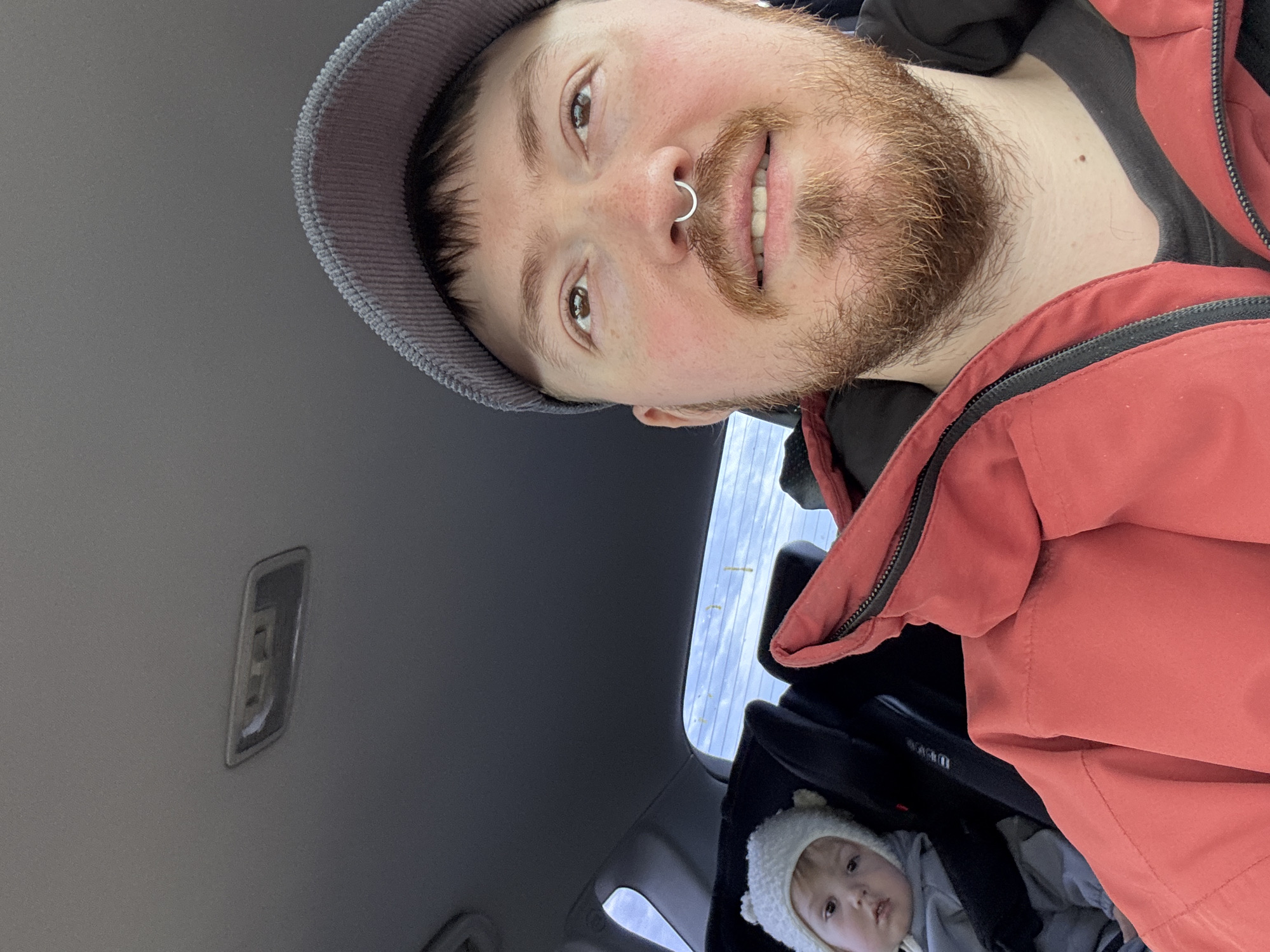 A selfie taken inside a car. A fair-skinned adult male with a reddish beard, wearing a gray hat and a red jacket, looks down at the camera with a slight smile. The man has a silver nose ring. A child wearing a white knit hat with bear ears sits in a black car seat behind the man. The car's interior is gray, and the car's ceiling light is visible. The location appears to be in an automobile; the city cannot be determined based on the visual information provided.